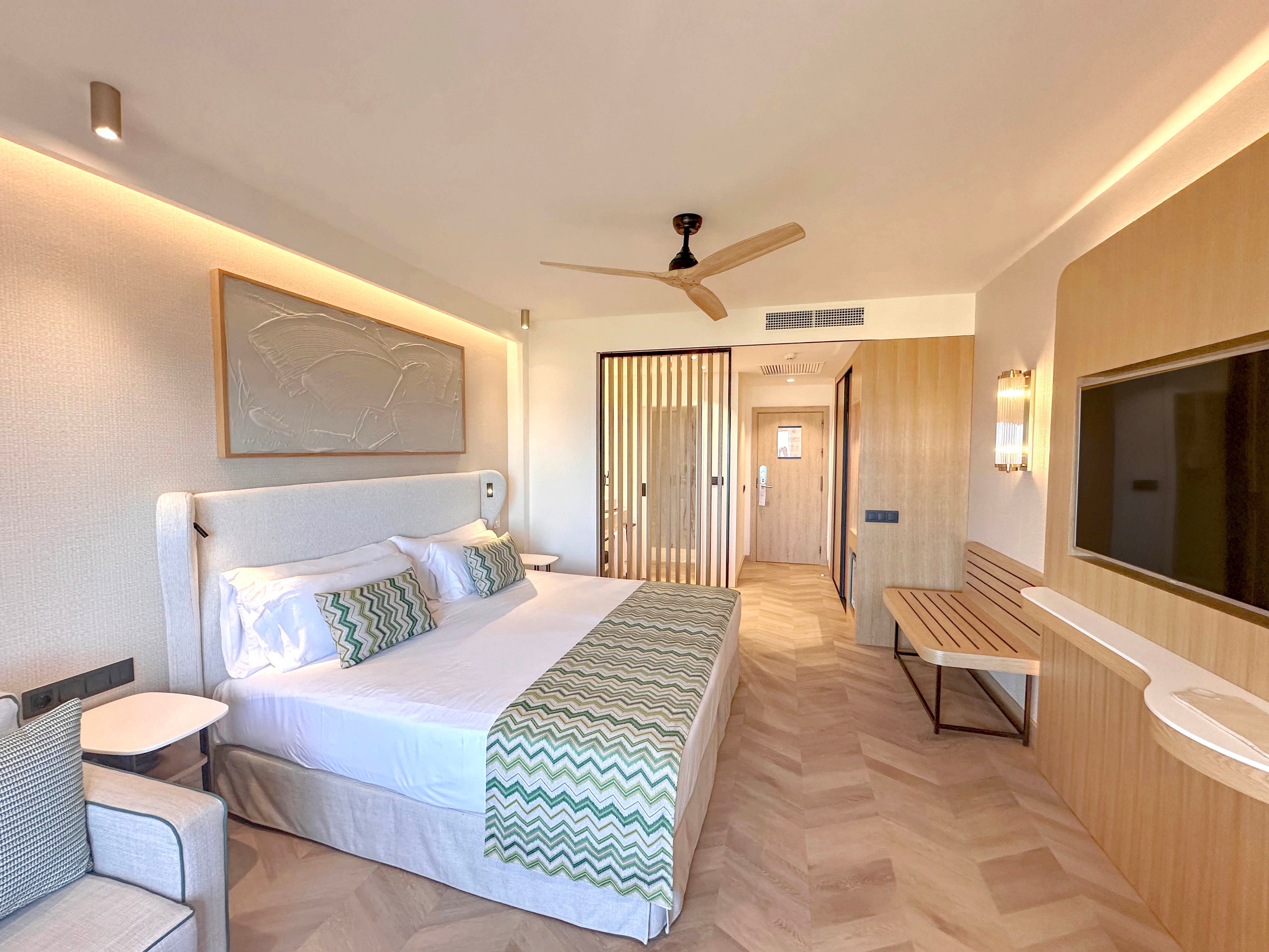A contemporary hotel bedroom features a large bed with white linens and patterned green accent pillows and runner. Light wood paneling and furniture create a warm, minimalist aesthetic throughout the room. A ceiling fan, wall-mounted television, and built-in desk area emphasize comfort and functionality. Soft lighting and neutral tones contribute to a calm, inviting atmosphere.