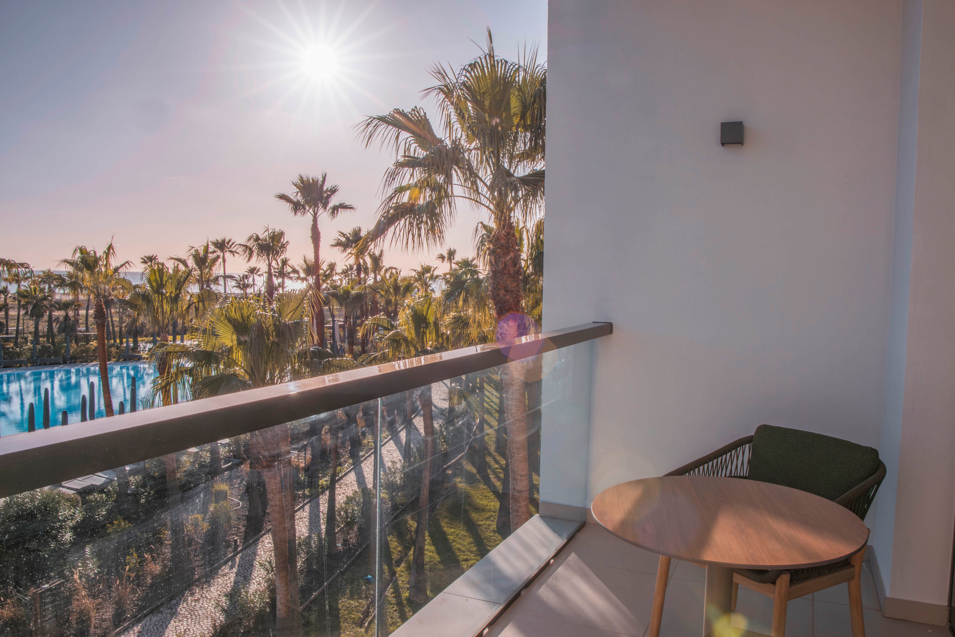 A modern balcony with a round wooden table and chair faces a lush outdoor scene filled with tall palm trees and a sparkling swimming pool. The sun shines brightly, casting warm light and creating a relaxed, vacation-like atmosphere. The glass railing offers an unobstructed view of the tropical landscape, emphasizing the serene and inviting setting. No visible text or numbers are present in the image.