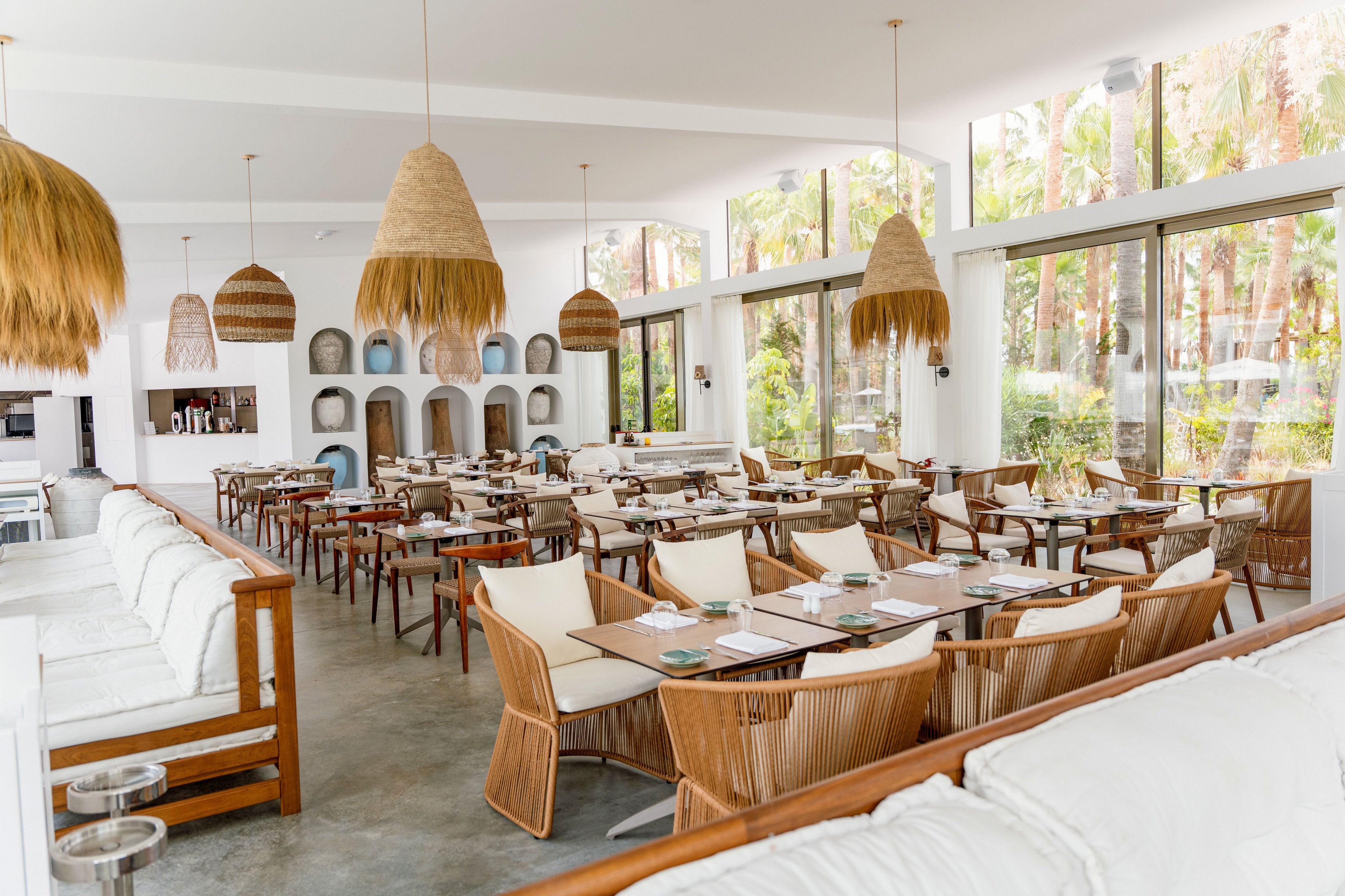 A spacious, modern restaurant interior features rows of wooden tables and wicker chairs arranged neatly. Large floor-to-ceiling windows let in abundant natural light and reveal lush greenery outside. White walls, cushioned bench seating, and woven pendant lights create a relaxed, coastal atmosphere. The space appears ready for guests, with place settings arranged on each table.