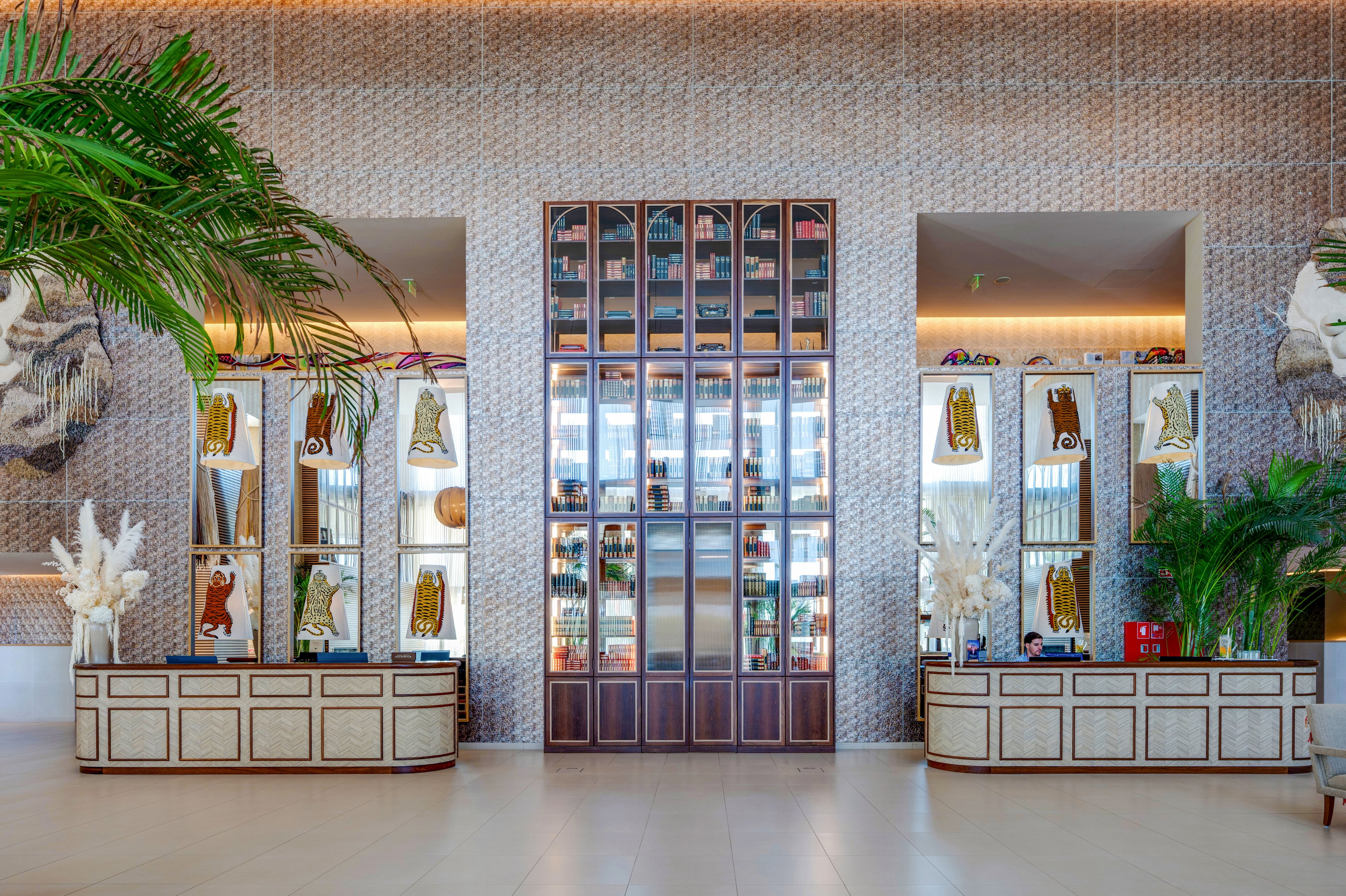 A contemporary hotel lobby features two reception desks flanking a tall central shelving unit filled with bottles and decorative objects. The walls are covered in a patterned design, complemented by framed artwork and illuminated display cabinets. Large green plants and white dried floral arrangements add a natural touch to the bright, open space. Warm ambient lighting and clean lines create an inviting, upscale atmosphere.