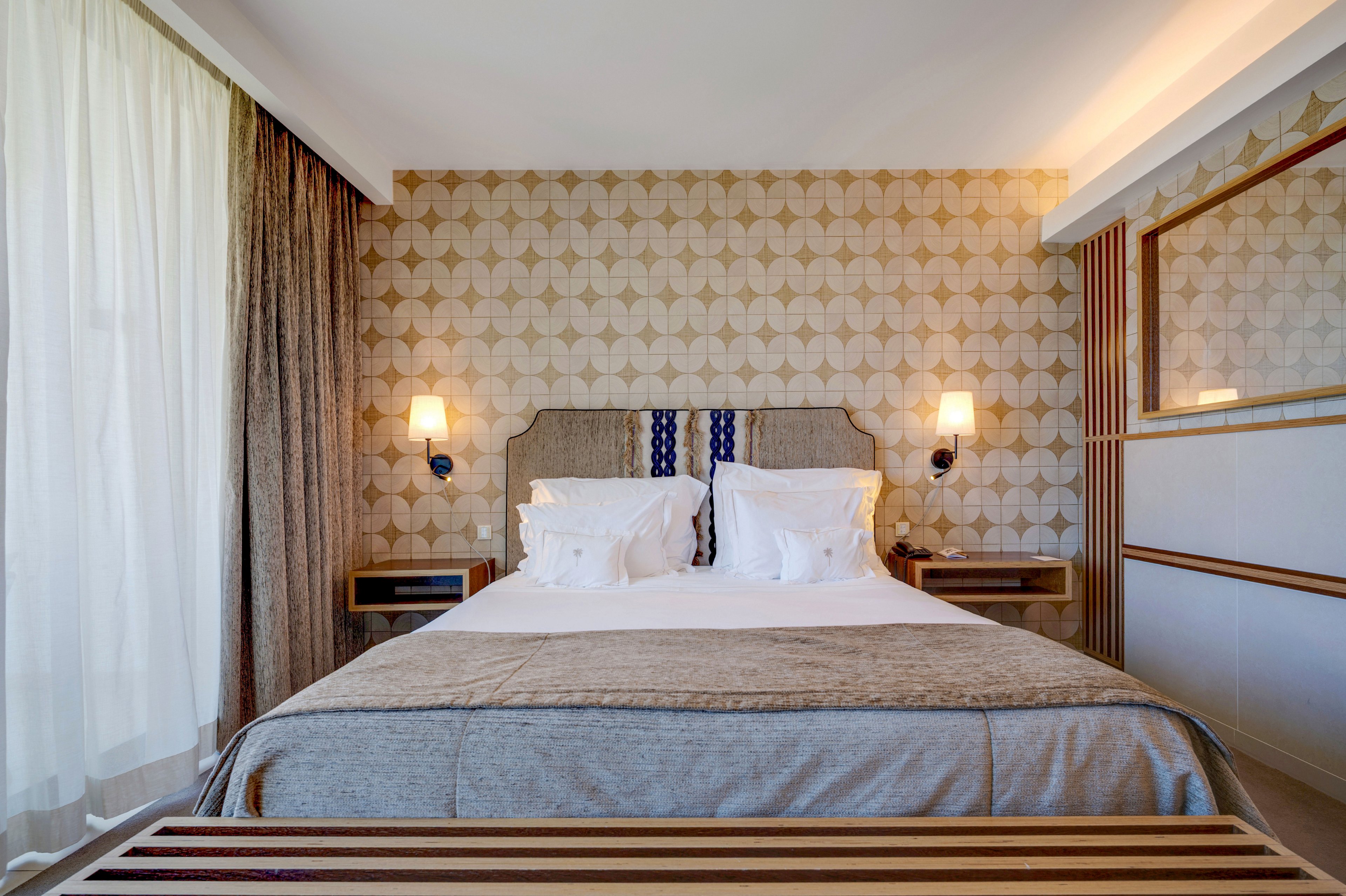 A neatly made double bed is centered in a modern hotel bedroom. The room features a geometric patterned accent wall, soft neutral bedding, and two wall-mounted lamps on either side of the headboard. Floor-to-ceiling curtains allow natural light to filter into the space, creating a bright and inviting atmosphere. A wooden bench sits at the foot of the bed, adding a touch of contemporary design.