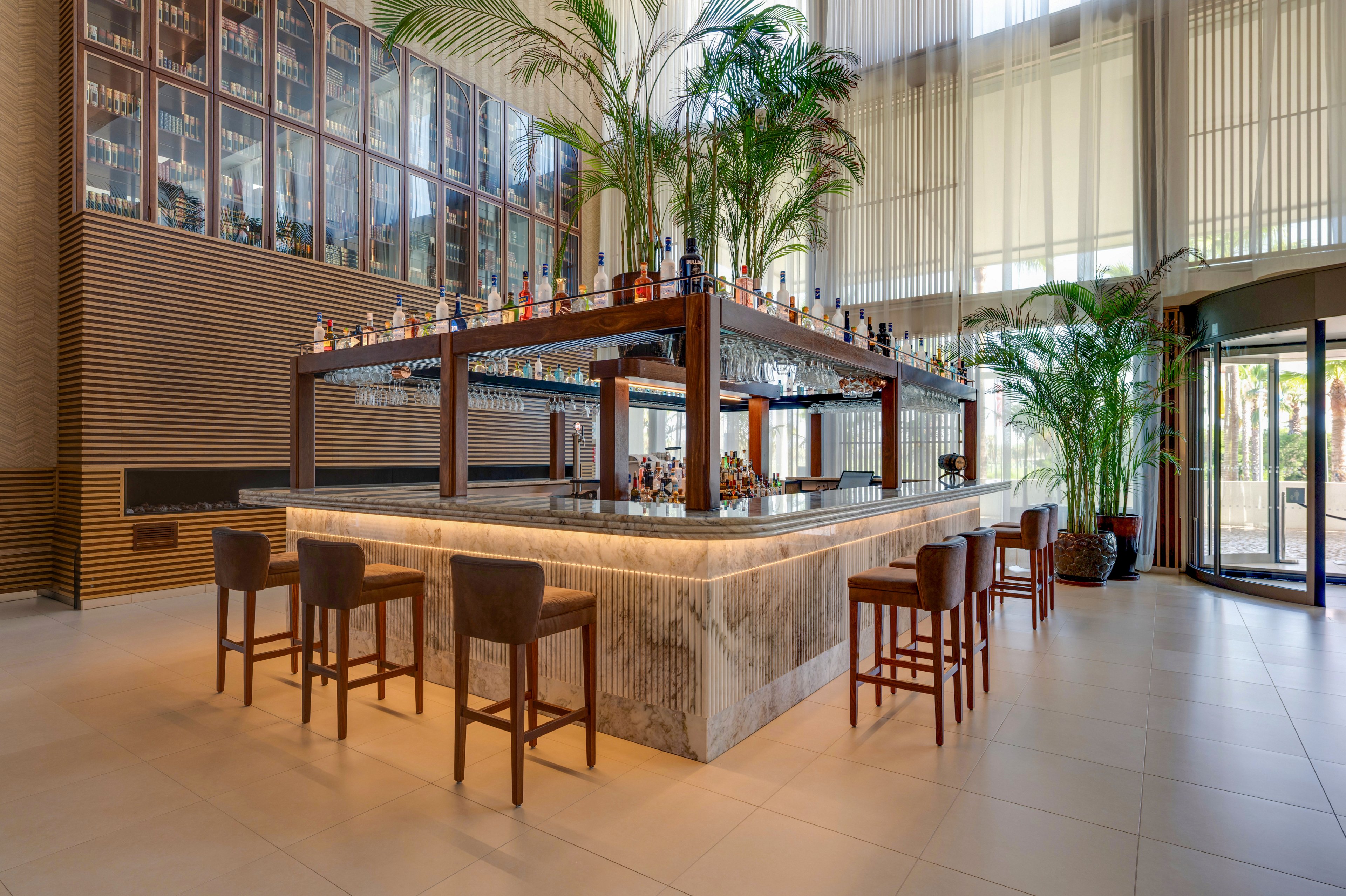 A contemporary bar with a large square island counter stands in the center of a spacious lobby. High ceilings with tall windows allow natural light to flood the room, highlighting the marble bar surface and warm wood accents. Shelves above the counter display numerous bottles and hanging glassware, while tall green plants add a tropical touch. Several brown leather bar stools surround the counter, creating an inviting setting for guests.