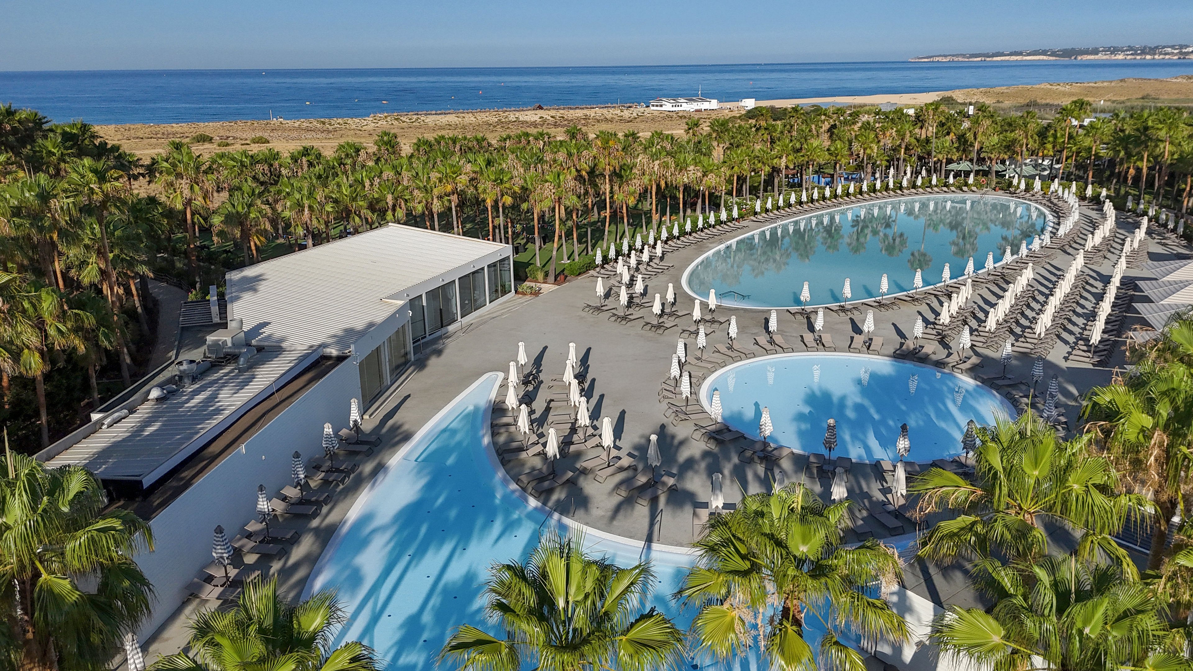 An aerial view showcases a modern coastal resort featuring two large circular swimming pools surrounded by palm trees. Numerous sun loungers and umbrellas are neatly arranged around the pools, suggesting a relaxing vacation atmosphere. A contemporary white building sits adjacent to the pool area, likely serving as a lounge or restaurant. In the background, a sandy shoreline and calm blue sea stretch toward the horizon under clear skies.