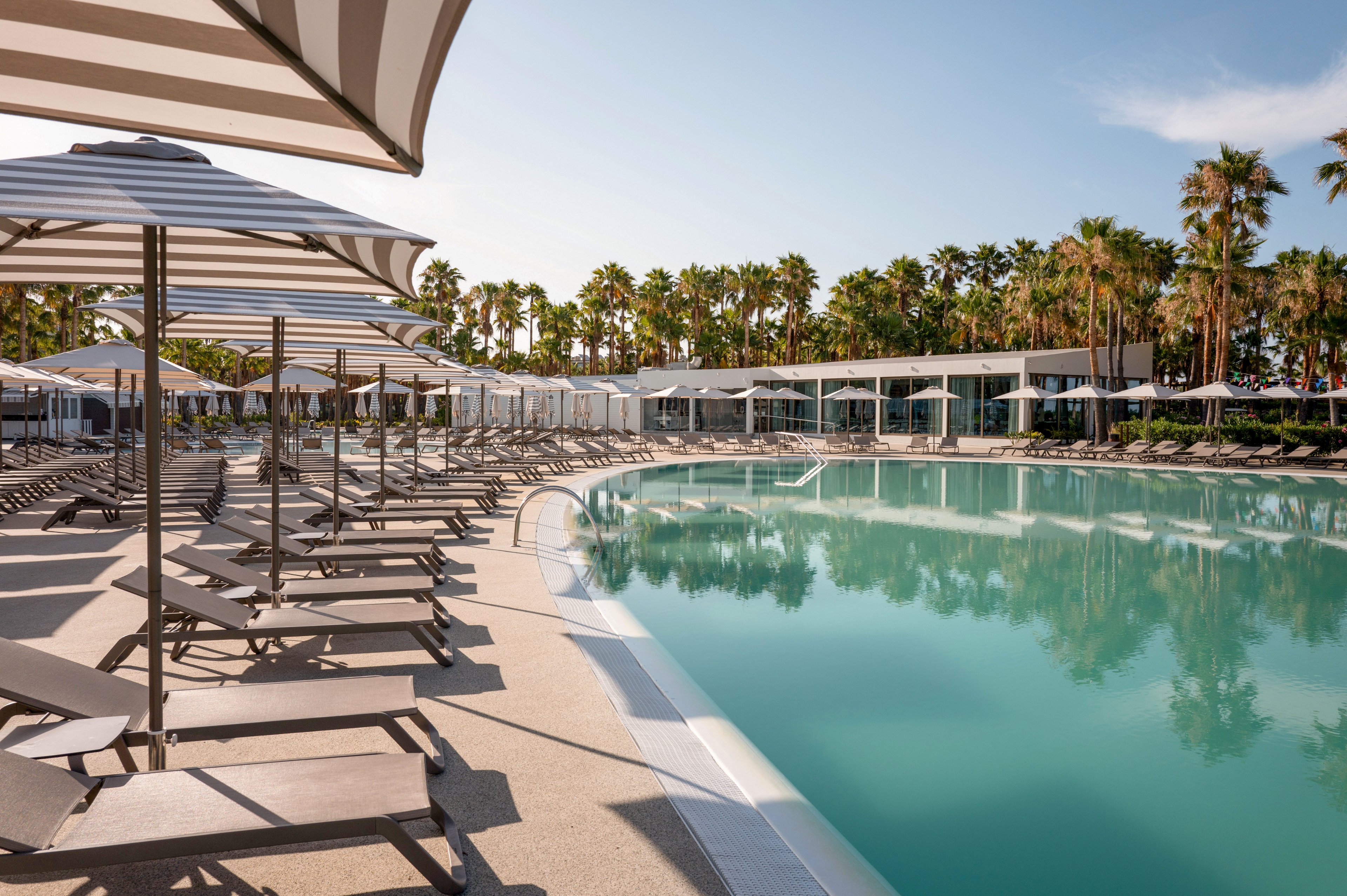 A large, curved swimming pool is surrounded by rows of empty sun loungers and striped parasols at a modern resort. The scene is set on a bright, clear day with calm turquoise water reflecting the sky. In the background, a line of tall palm trees frames sleek white buildings with large windows. The overall atmosphere is tranquil and inviting, suggesting a relaxing vacation destination.
