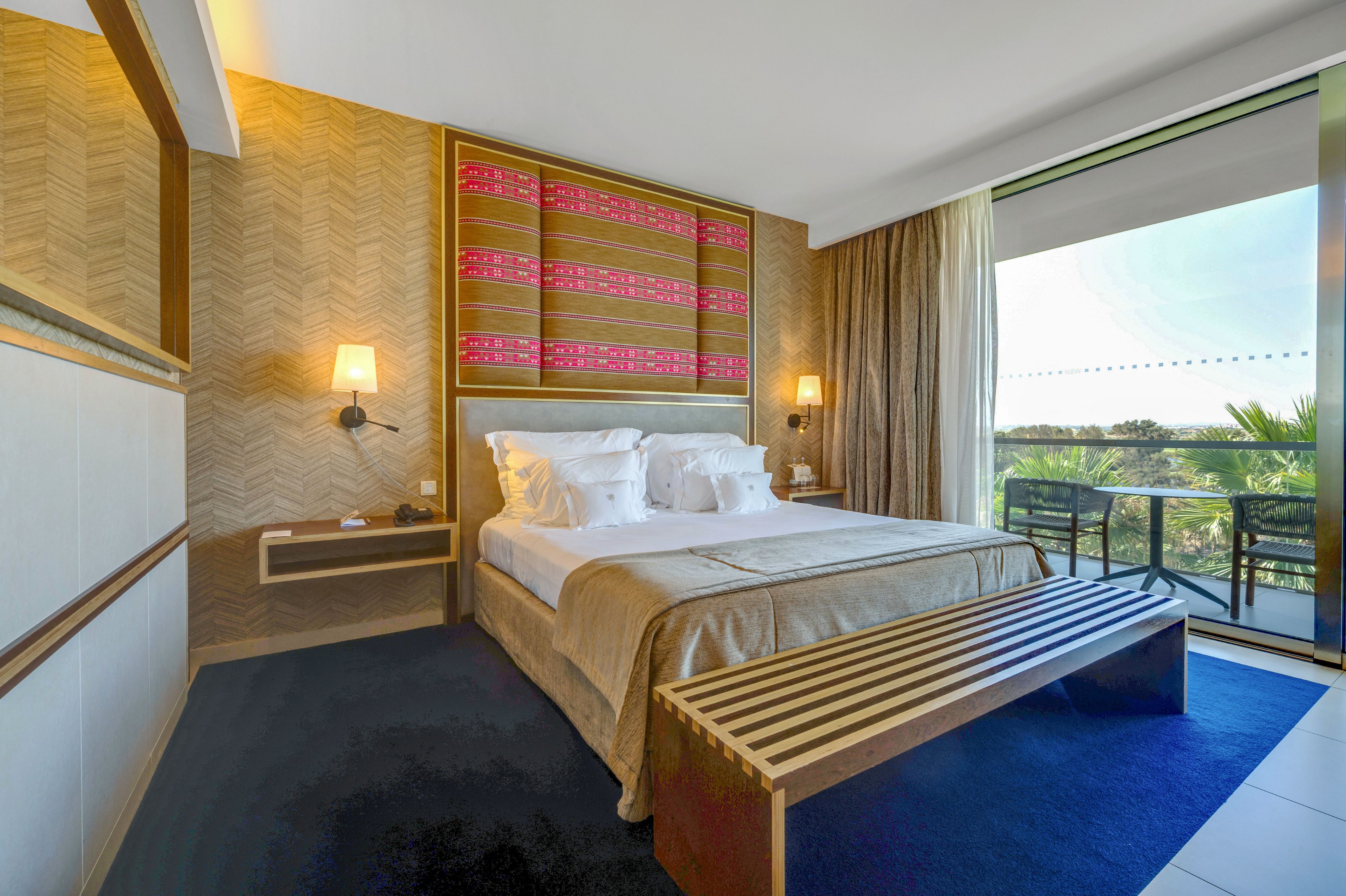 A contemporary hotel bedroom features a neatly made double bed with crisp white linens and a beige bedspread. Warm wooden accents, patterned wall panels, and soft lighting create a cozy, upscale atmosphere. A slatted wooden bench sits at the foot of the bed on a deep blue rug. Floor-to-ceiling curtains open to a balcony with glass railing, overlooking trees and a sunny outdoor landscape.