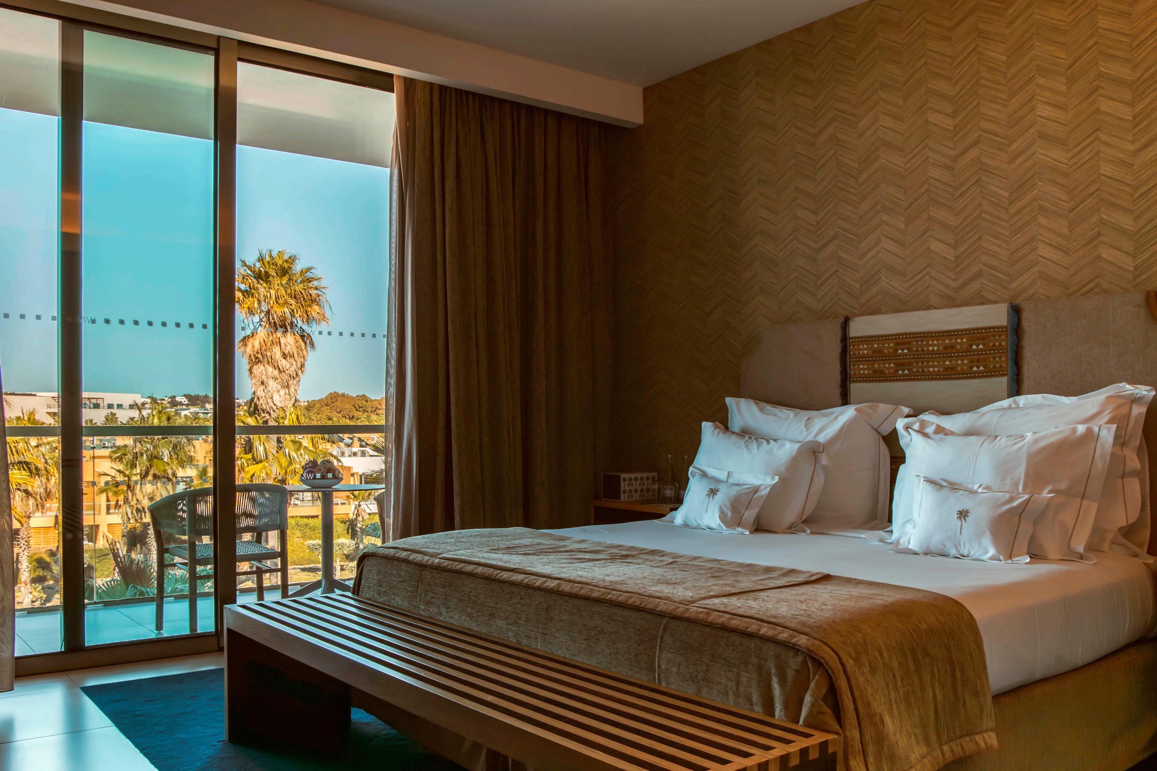 A contemporary hotel room features a neatly made bed with multiple pillows and a cushioned bench at the foot. Large glass doors open to a balcony overlooking palm trees and a pool area, letting in natural daylight. The room has a warm, neutral palette with textured walls and elegant decor, creating a relaxing atmosphere.