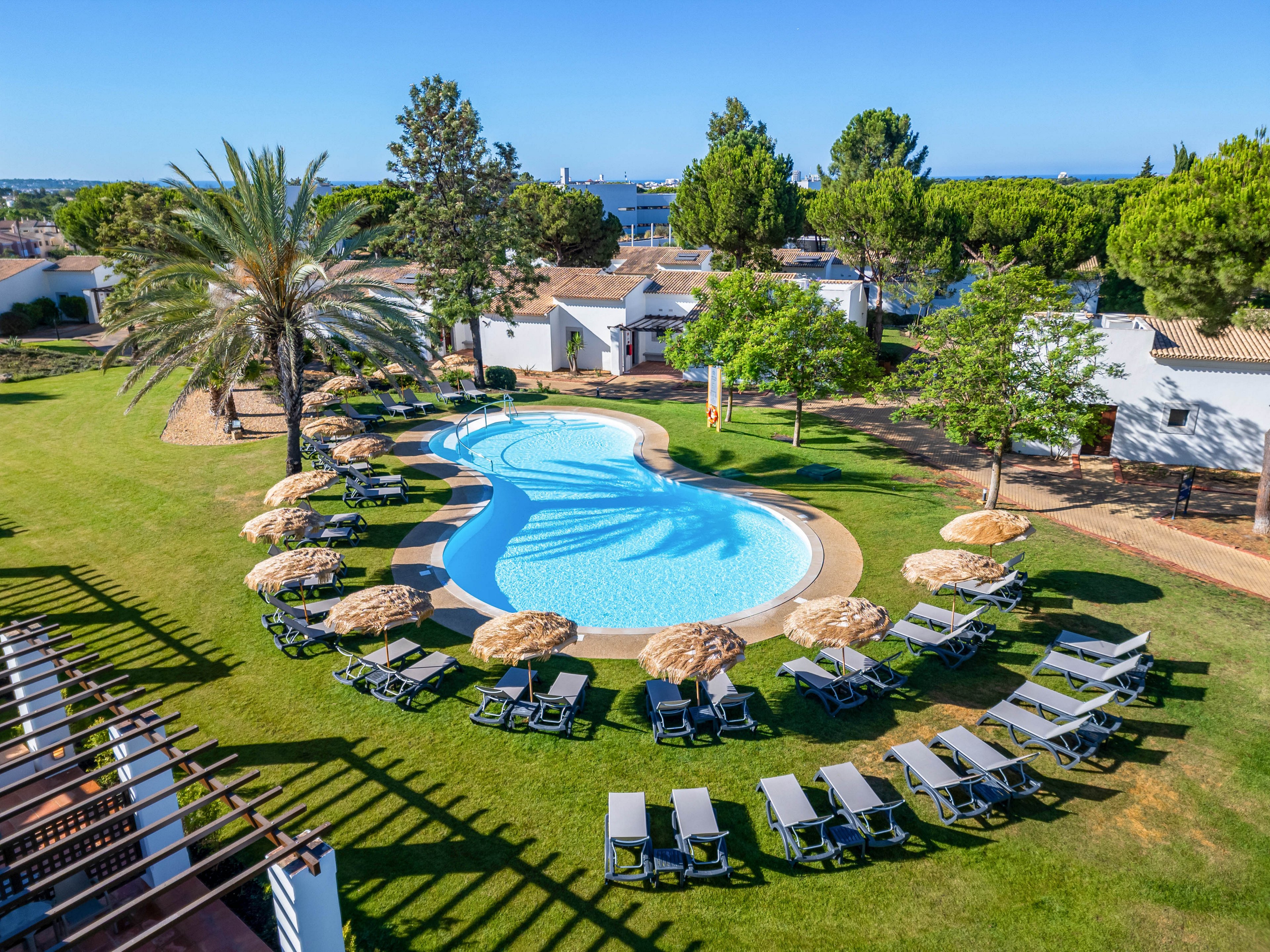 An outdoor resort swimming pool curves through a neatly manicured lawn surrounded by sun loungers and straw parasols. White low-rise buildings and tall green trees frame the pool area, creating a relaxed vacation setting. The bright blue water contrasts with the vivid green grass under clear, sunny skies. The scene suggests a peaceful holiday destination with ample space for guests to relax.