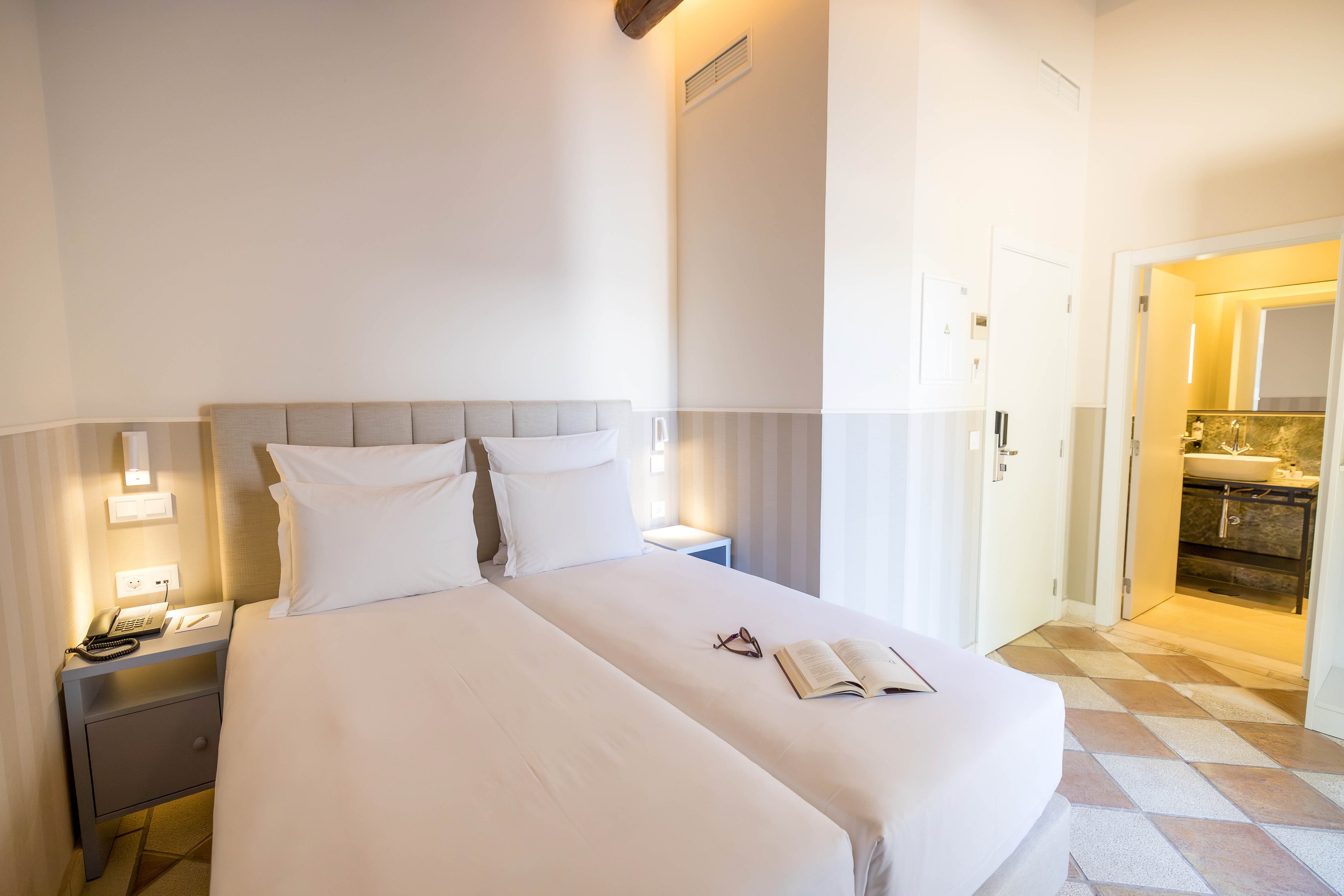 A neatly made double bed with white linens stands in a bright, modern hotel room. A bedside table with a phone and wall-mounted lights frames the bed, while an open book and eyeglasses rest on the duvet. The room features soft neutral colors, striped lower walls, and a tiled floor leading toward an illuminated ensuite bathroom. The bathroom area includes a sink, mirror, and storage, contributing to a clean and inviting atmosphere.