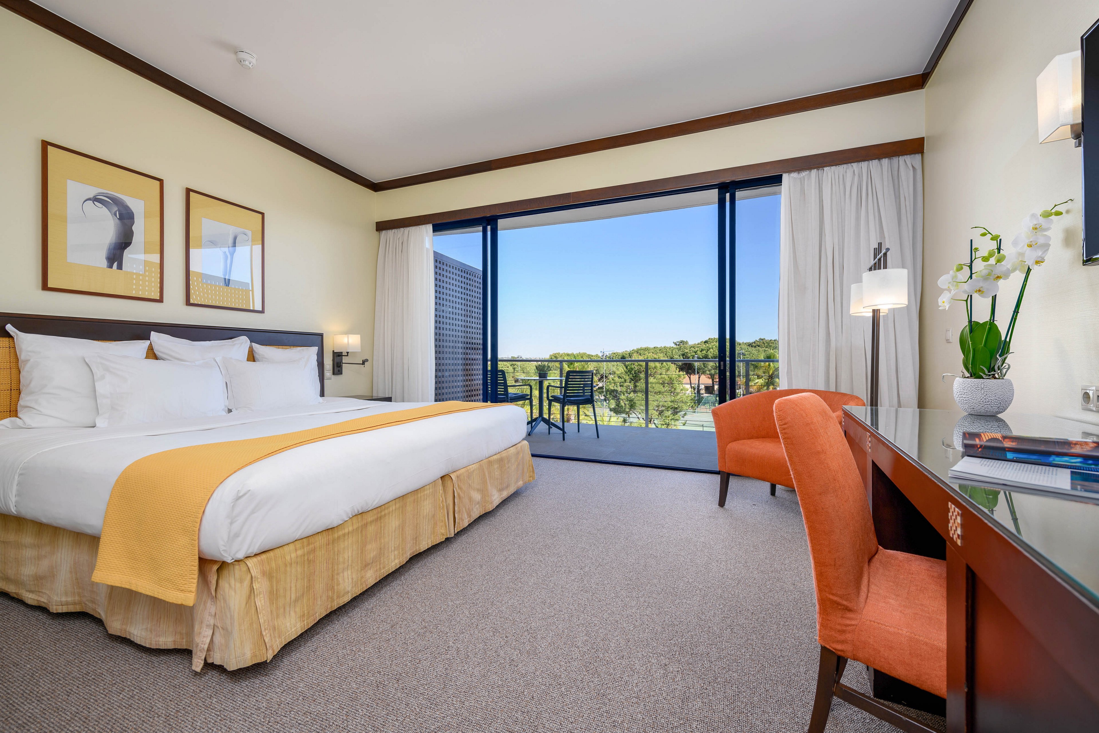 A spacious hotel bedroom features a large neatly made bed with white linens and a yellow throw. Floor-to-ceiling glass doors open to a balcony with chairs overlooking a green landscape and blue sky. The room includes framed artwork above the bed, an orange upholstered chair, a glass-topped desk, and decorative lighting. Neutral walls and carpeting create a clean, contemporary atmosphere with natural light filling the space.