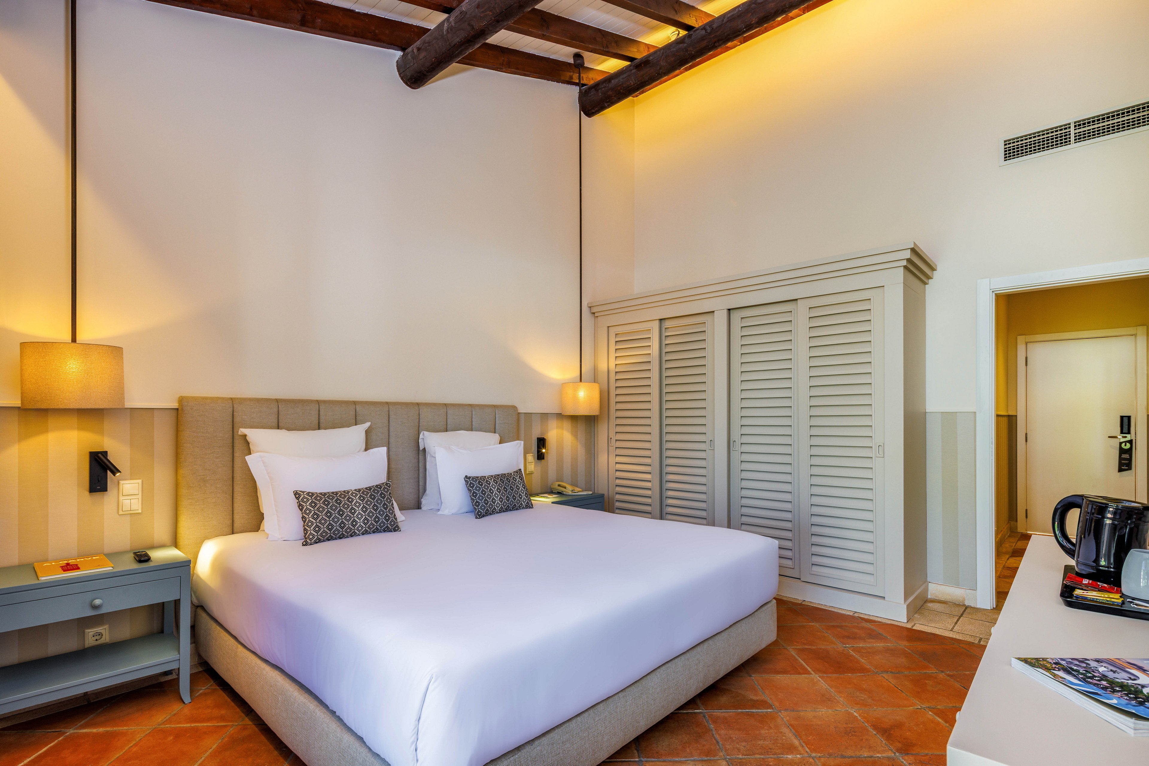 Bright, inviting hotel room featuring a large bed with white linens and decorative pillows. Warm lighting, exposed wooden ceiling beams, and terracotta floor tiles create a cozy atmosphere. Built-in wardrobe doors, bedside tables, and wall-mounted reading lamps add functional comfort. A kettle and cups on the side counter suggest in-room refreshments, while the doorway leads to an adjacent area.