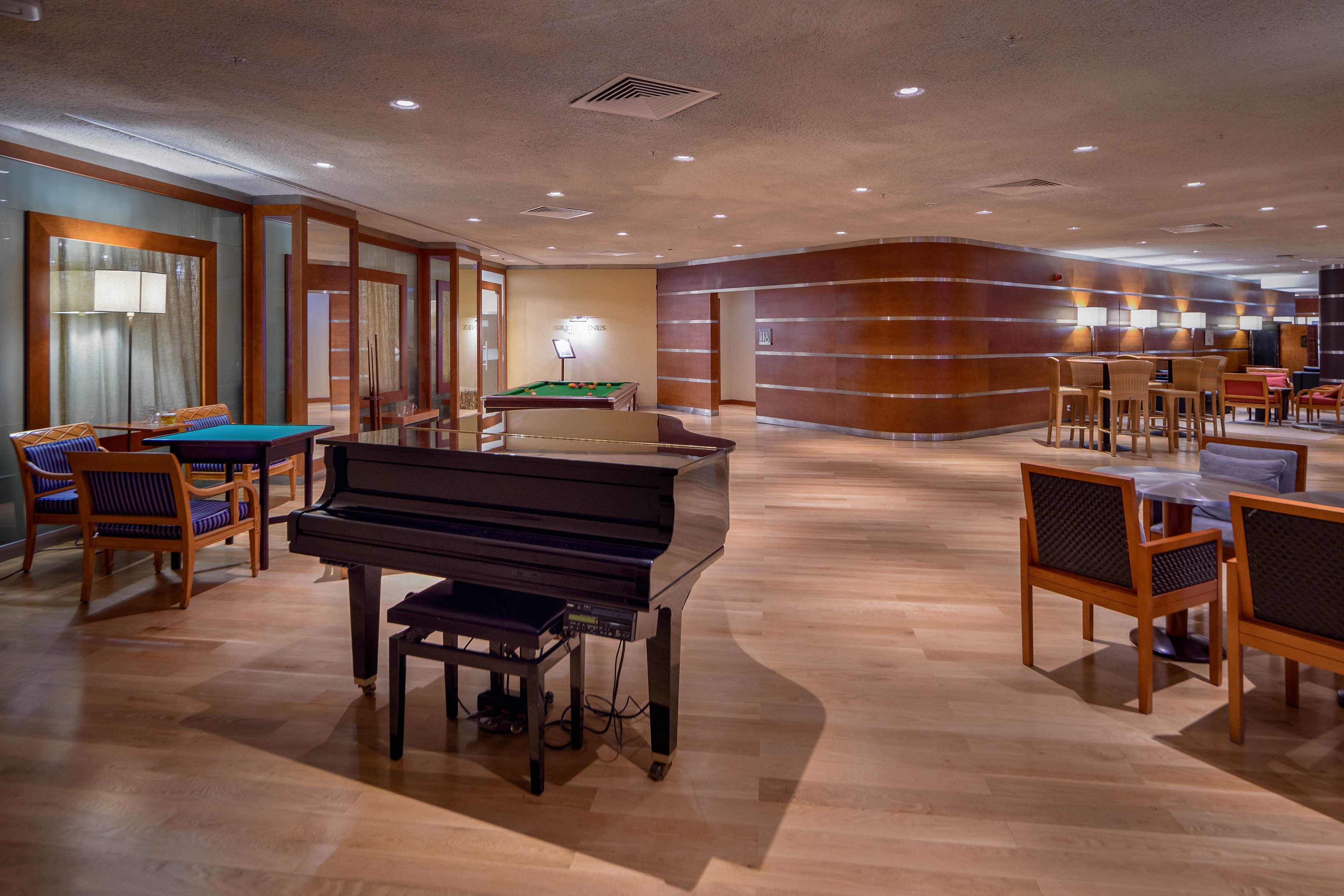 A polished black grand piano stands prominently in a spacious lounge area. The room features wooden flooring, warm wood-paneled walls, and contemporary seating with tables and chairs arranged for socializing or relaxation. Soft ambient lighting from ceiling fixtures and floor lamps creates a cozy, upscale atmosphere. Glass partitions and a bar area in the background suggest a refined hotel or club setting designed for entertainment and comfort.
