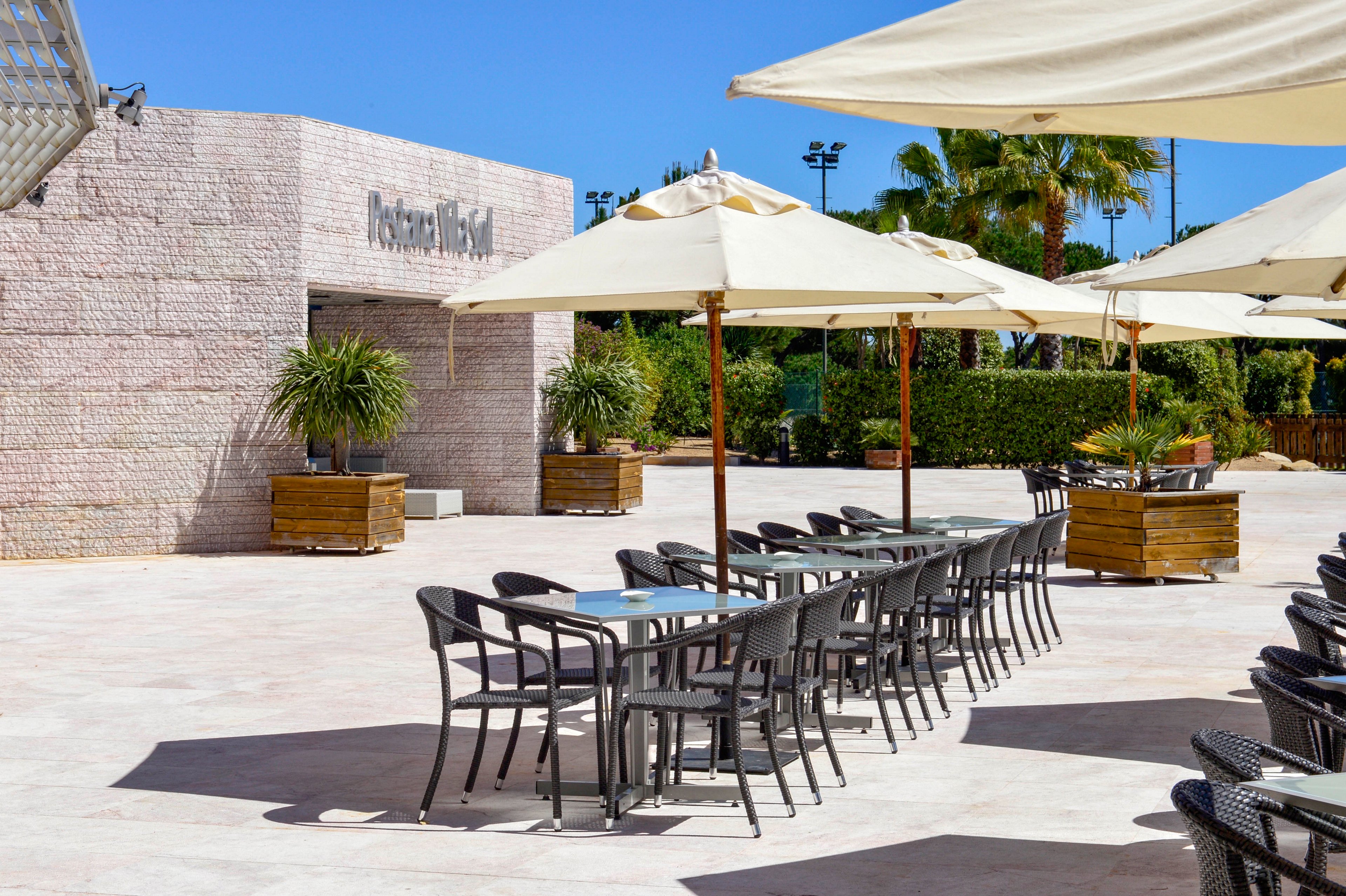 An empty outdoor restaurant terrace features rows of black chairs and glass-top tables under large cream-colored sun umbrellas. The setting is a bright, sunny day with clear blue sky and strong shadows on the tiled floor. Potted green plants in wooden planters line the space, and a stone wall with visible text marks the restaurant entrance. Palm trees and manicured hedges frame the background, creating a relaxed vacation atmosphere.