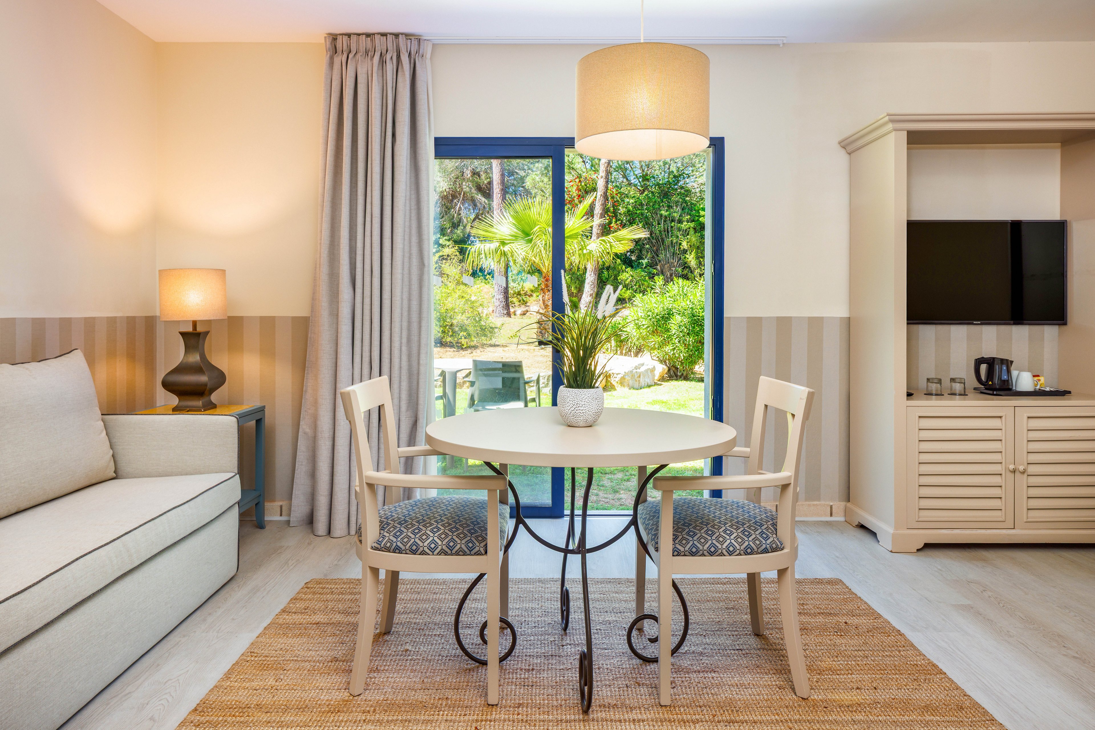 A cozy hotel room features a round dining table with two chairs placed on a woven rug. A sofa and side table with a lit lamp sit to the left, while a cabinet with a flat-screen TV and coffee station is on the right. Large glass doors open to a lush garden with palm trees and outdoor seating. Soft neutral tones and warm lighting create a welcoming, relaxed atmosphere.