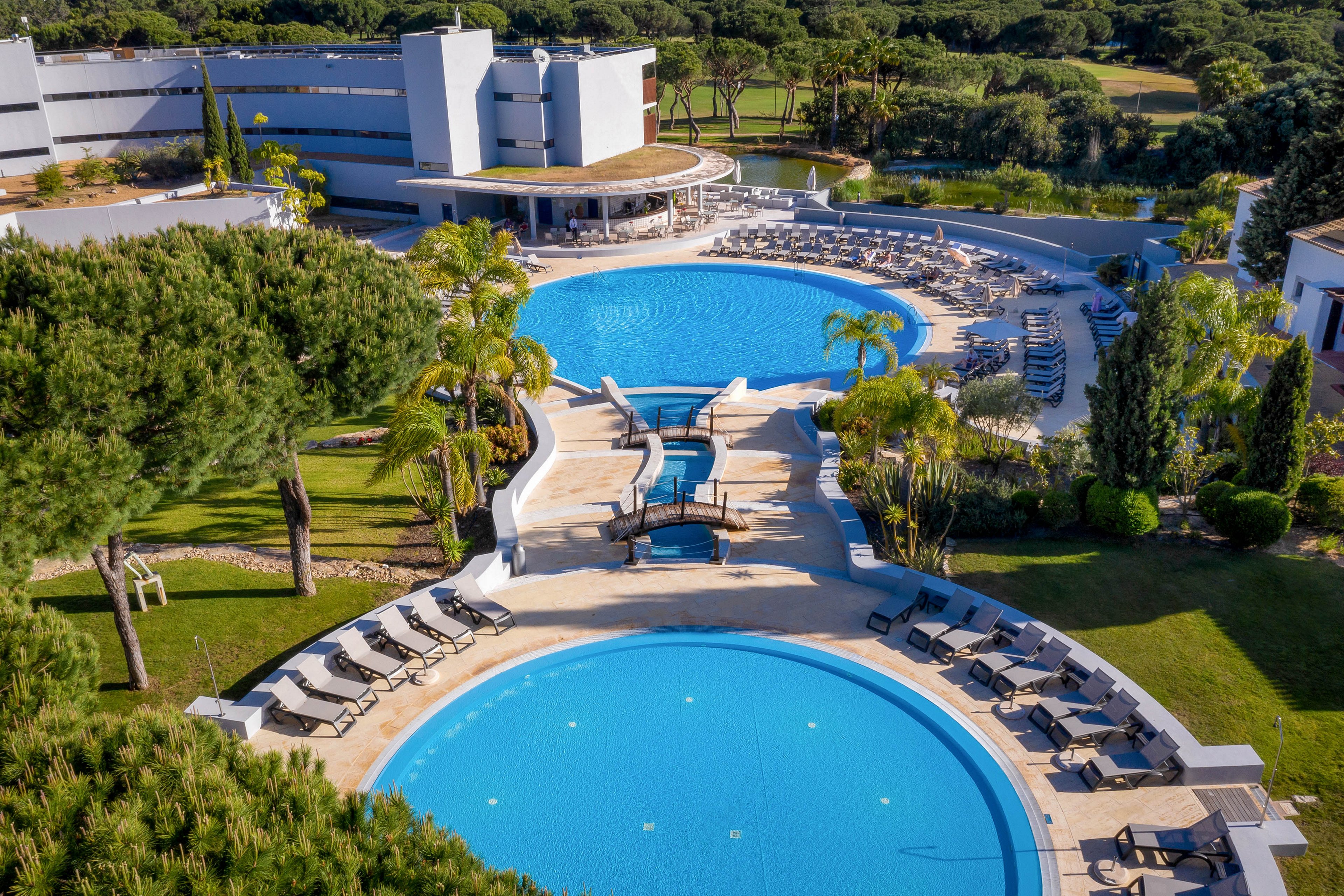 A modern resort hotel features two large circular outdoor swimming pools surrounded by sun loungers and umbrellas. The pools sit on a landscaped terrace with palm trees, manicured lawns, and stone pathways. A curved poolside bar or pavilion connects to the main building, which overlooks a lush forested area in the background. The bright blue water and greenery create a relaxing vacation atmosphere under clear skies.
