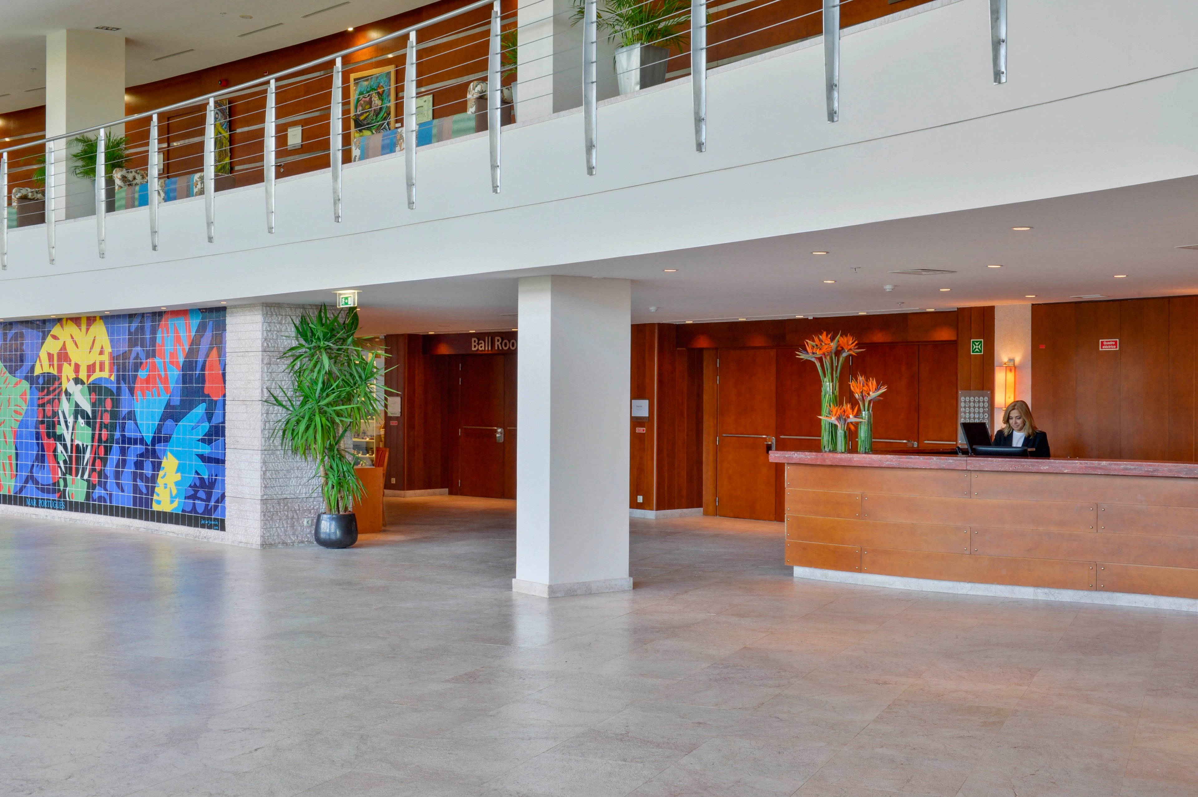 Spacious hotel lobby interior features a wooden reception desk staffed by a person. The setting includes light tiled flooring, white walls, and warm wooden paneling with doors behind the desk. A colorful abstract mural and large potted plants add visual interest along one wall. An upper mezzanine with metal railings overlooks the lobby, creating an open and contemporary atmosphere.