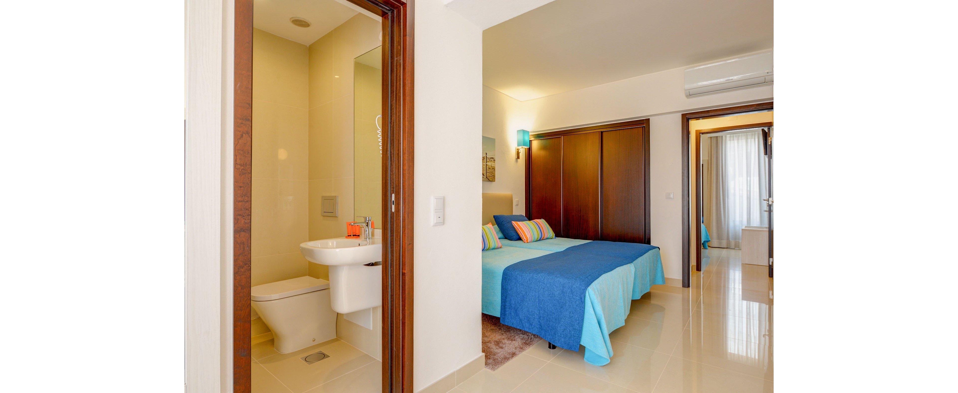 A contemporary hotel-style bedroom features a double bed with blue bedding and colorful striped pillows. The room has glossy tiled floors, wooden wardrobe doors, and a wall-mounted light casting a warm glow. An open doorway reveals a compact ensuite bathroom with a white sink and toilet. Natural light enters through a window or balcony door in the background, enhancing the clean and inviting atmosphere.