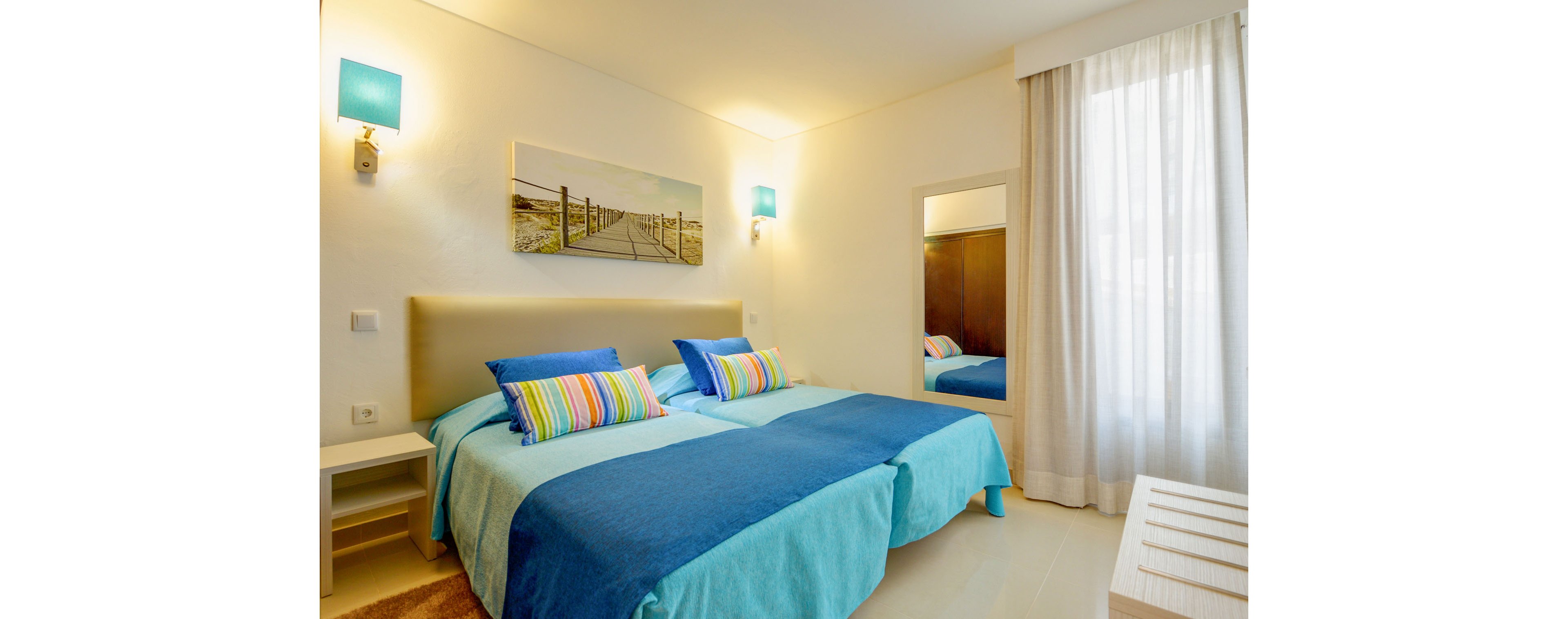 A contemporary bedroom features two twin beds dressed in turquoise and blue bedding. Colorful striped pillows add a playful accent against the neutral headboard and walls. Soft lighting from wall sconces and natural light from a sheer-curtained window create a warm, inviting atmosphere. A landscape artwork above the beds and minimal furniture complete the clean, modern look.