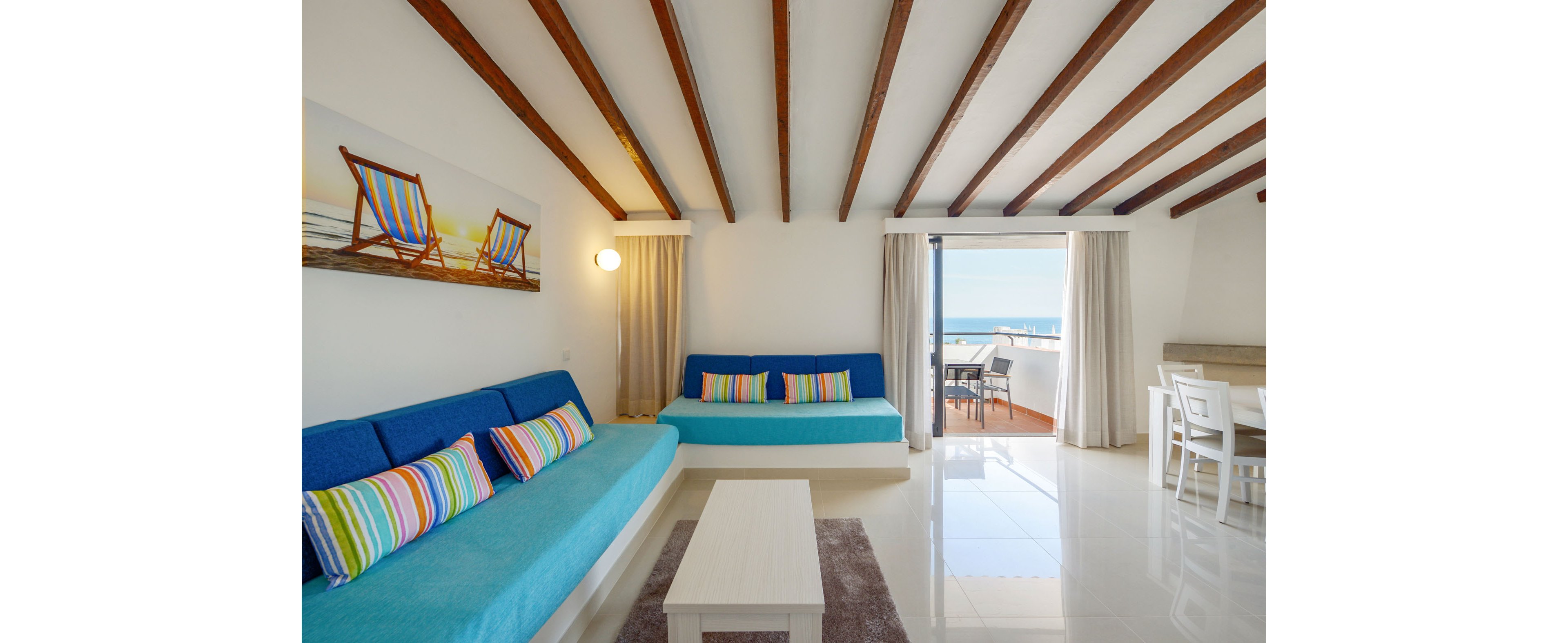 Sunny vacation-style living room with two blue sofas and colorful striped cushions. Wooden ceiling beams and light tiled floors create an airy, modern interior. A wall art print of deck chairs and a small coffee table sit in the foreground. Sliding glass doors open to a balcony with outdoor seating and an ocean view, while a compact dining area is visible to the right.