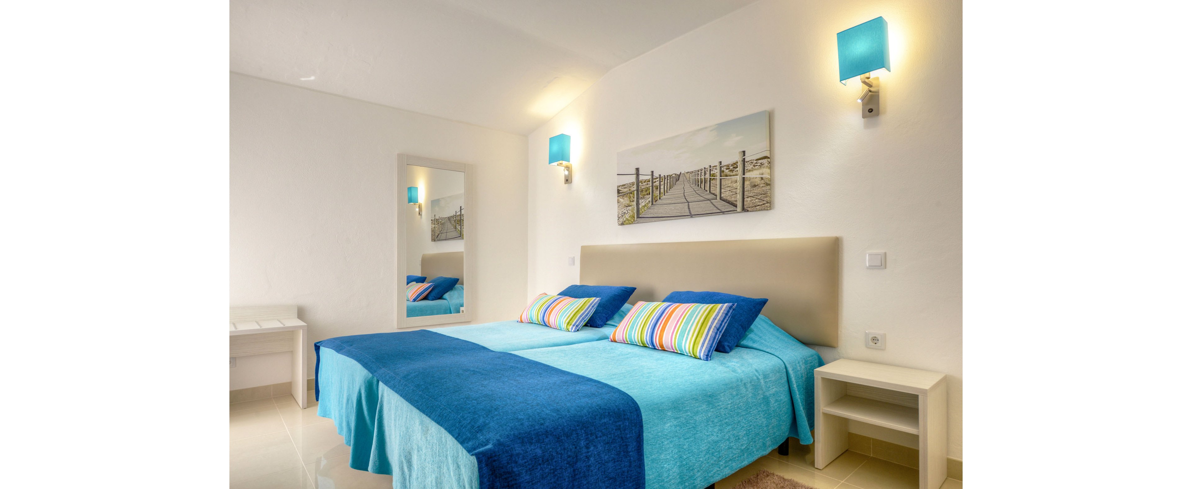 Modern bedroom interior featuring two twin beds with turquoise and deep blue bedding. Colorful striped pillows add a playful accent against the neutral headboard and white walls. Blue wall sconces provide soft lighting, complementing the coastal artwork above the bed. A wall mirror, bedside tables, and a small desk complete the clean, contemporary space.