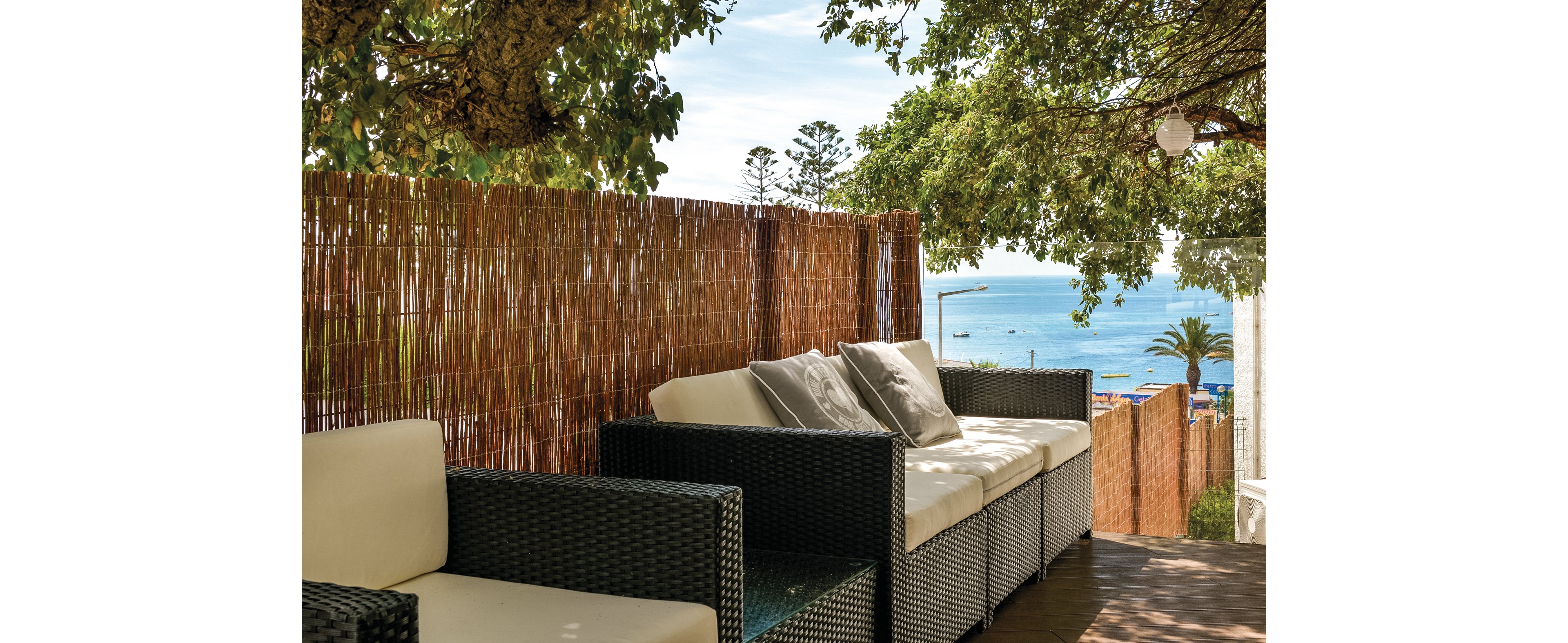 A serene outdoor patio featuring wicker furniture with white cushions, set against a backdrop of bamboo fencing. The setting overlooks a picturesque ocean view with boats and palm trees visible in the distance. The area is shaded by lush green trees, creating a tranquil and inviting atmosphere.