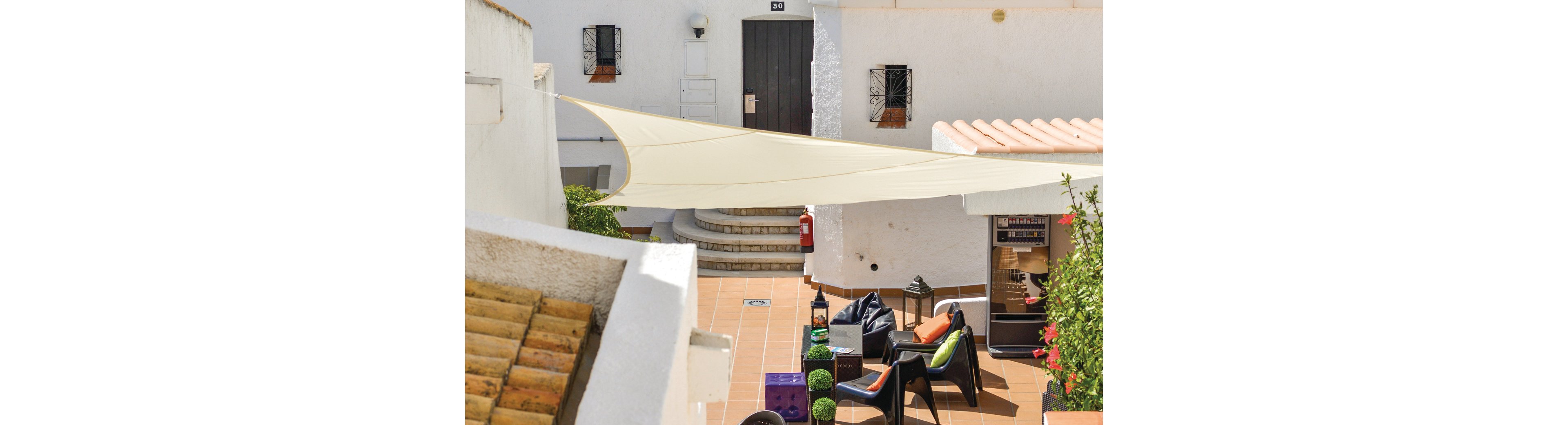 A bright and inviting courtyard featuring a white shade sail stretched over a tiled patio. The area includes modern outdoor seating with colorful cushions, potted plants, and a vending machine in the background. The white walls and terracotta roof tiles create a Mediterranean ambiance, complemented by natural light.