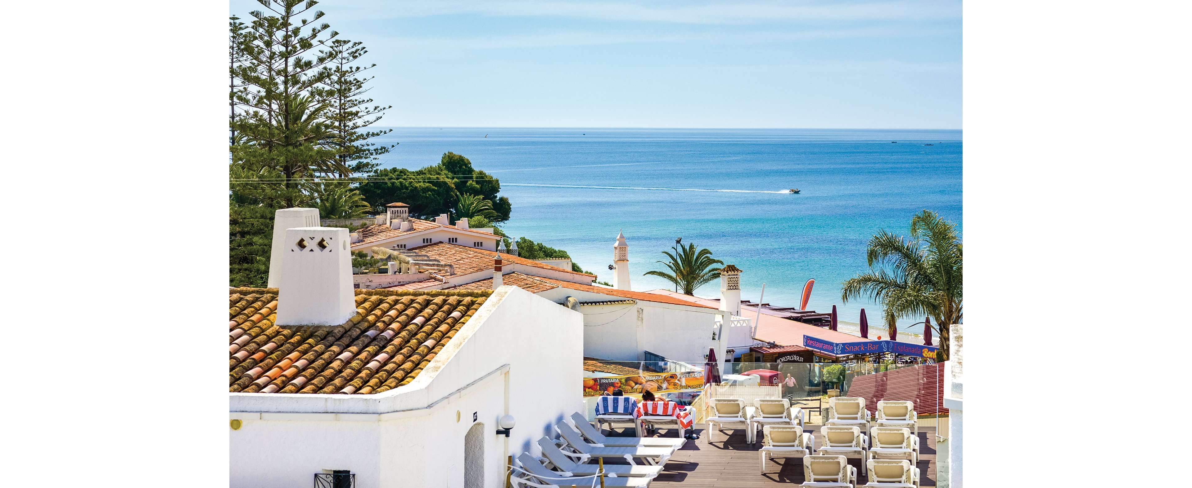A scenic coastal resort overlooking the ocean, featuring whitewashed buildings with terracotta roofs. The setting includes a terrace with multiple sun loungers arranged for relaxation. Palm trees and vibrant umbrellas add to the tropical ambiance, with clear blue waters visible in the background.