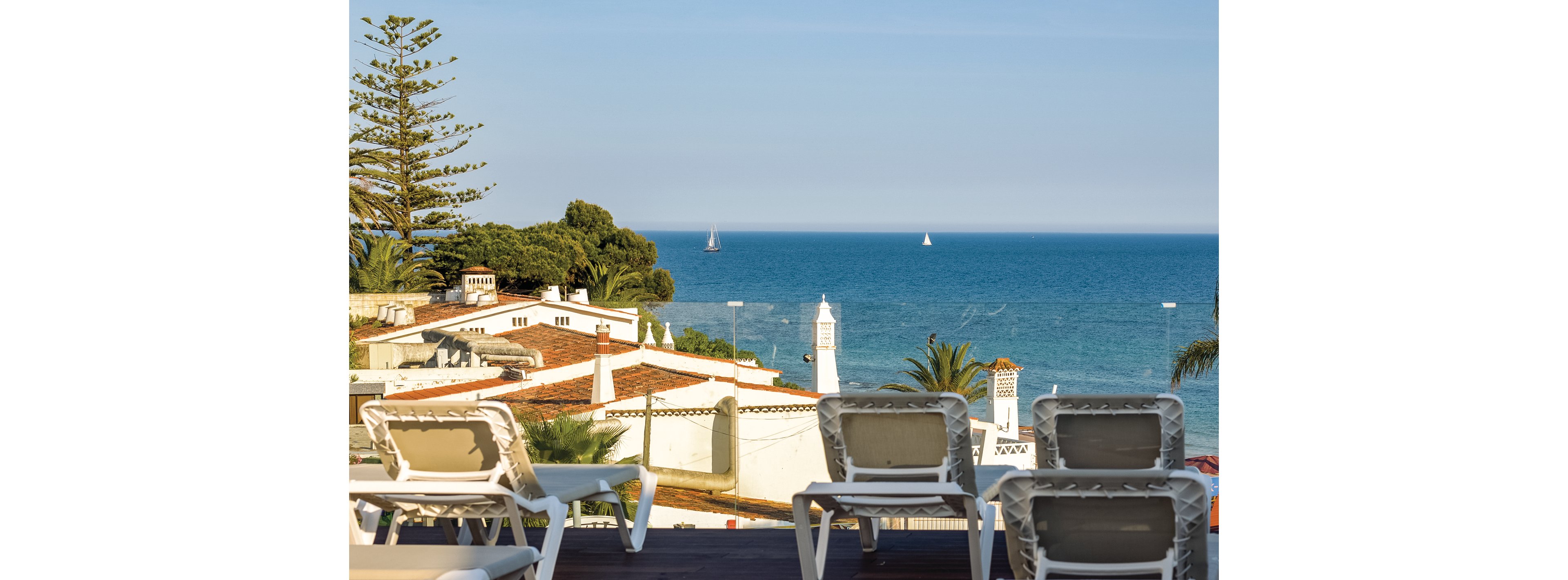 A serene coastal scene featuring white lounge chairs on a wooden deck overlooking rooftops and the blue ocean. Sailboats are visible in the distance, adding to the tranquil atmosphere. The setting is sunny with clear skies, surrounded by greenery and palm trees.