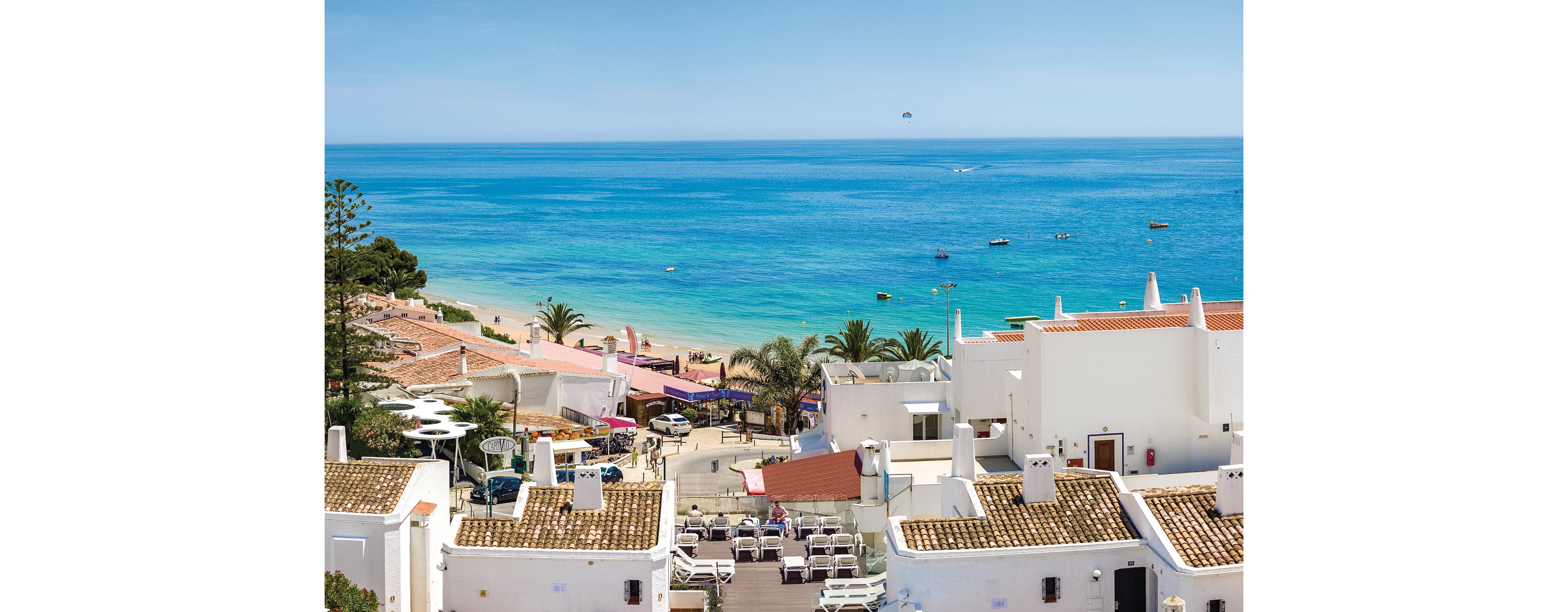 A picturesque coastal village featuring whitewashed buildings with terracotta rooftops overlooking a vibrant blue ocean. The setting includes palm trees, outdoor seating areas, and small boats scattered across the water. The scene evokes a tranquil and sunny Mediterranean atmosphere.