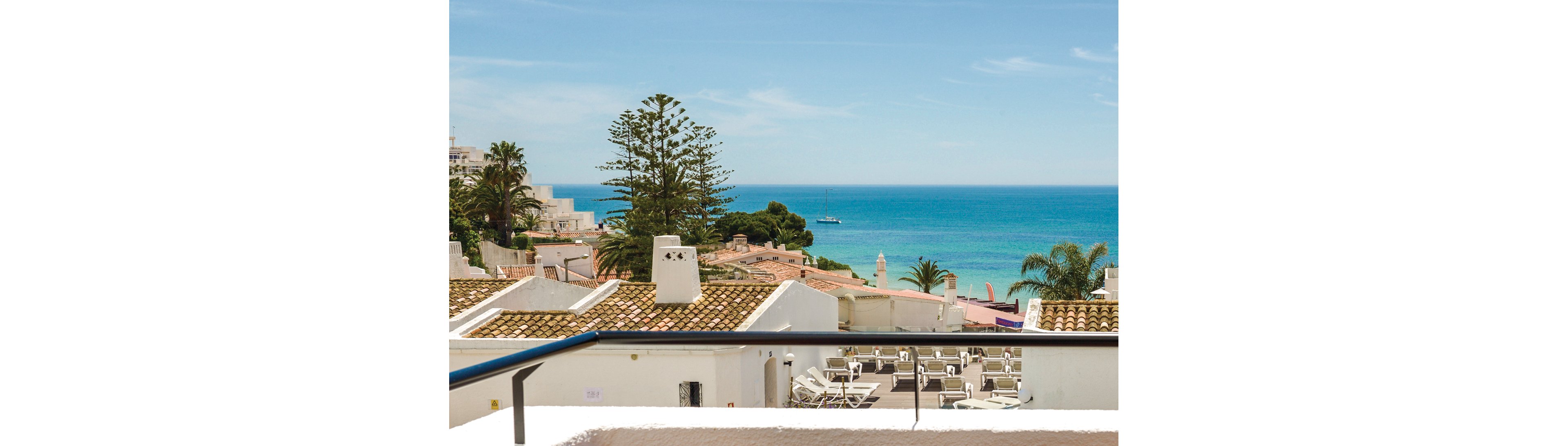 A picturesque coastal scene featuring white rooftops and a clear view of the ocean. The setting includes lounge chairs on a terrace, palm trees, and a sailboat in the distance. The bright blue sky and vibrant sea create a serene and inviting atmosphere.
