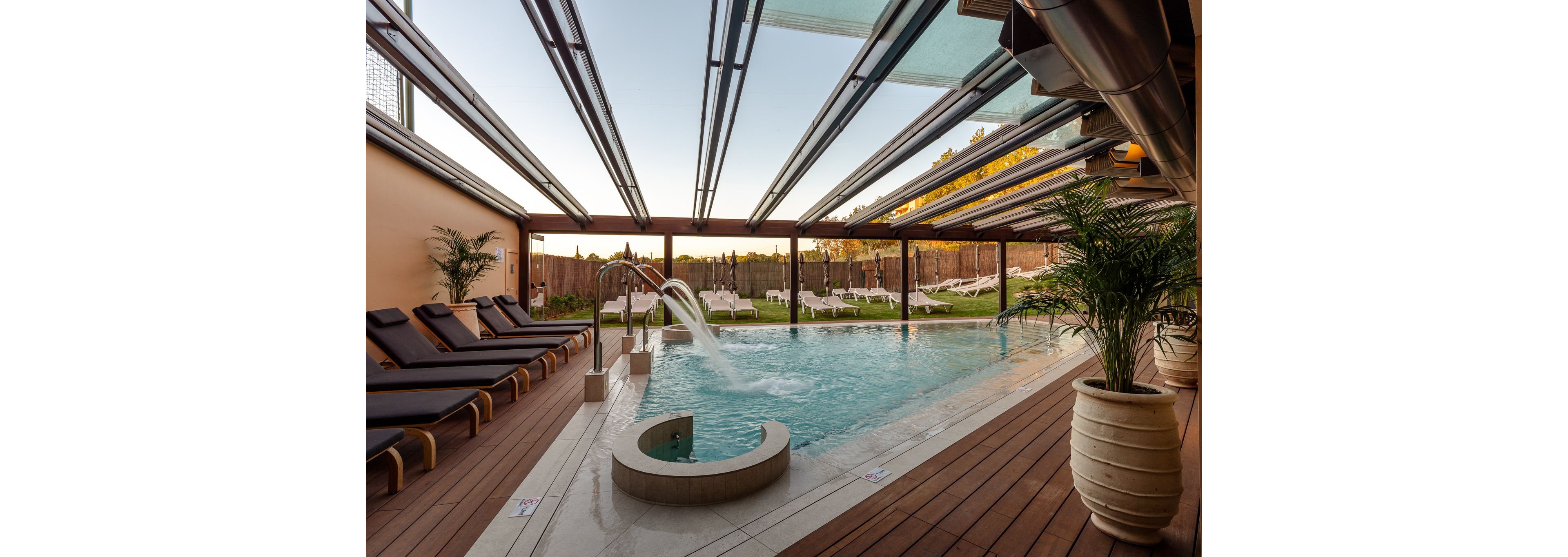 A contemporary outdoor swimming pool sits beneath a partially glass-covered structure. Wooden decking surrounds the pool, lined with cushioned lounge chairs on one side and a large potted plant in the foreground. Water flows from a curved metal fountain into the pool, adding a spa-like touch. In the background, white sun loungers and green lawn create a relaxing resort atmosphere at sunset.