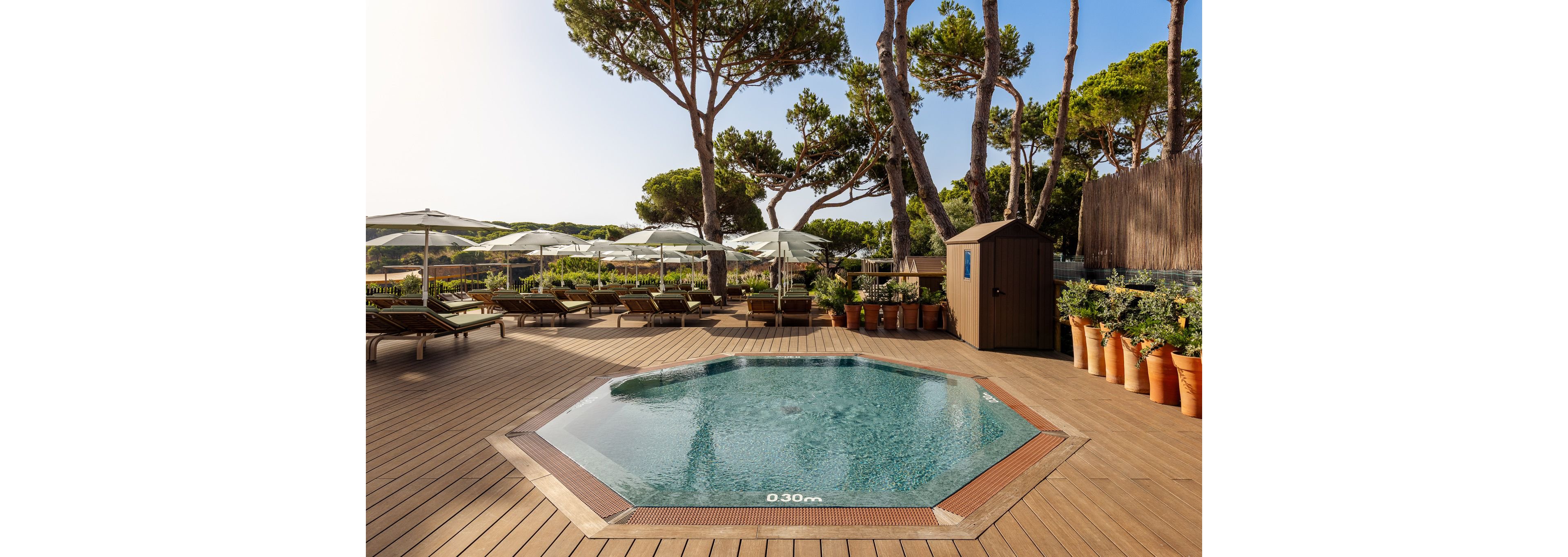 An octagonal spa pool sits in the center of a wooden deck in an outdoor relaxation area. The water is clear and calm, with the depth marking "0.30 m" visible on the pool edge. Surrounding the pool are sun loungers, white parasols, and potted plants, framed by tall pine trees. The scene suggests a tranquil resort or hotel terrace designed for leisure and comfort.