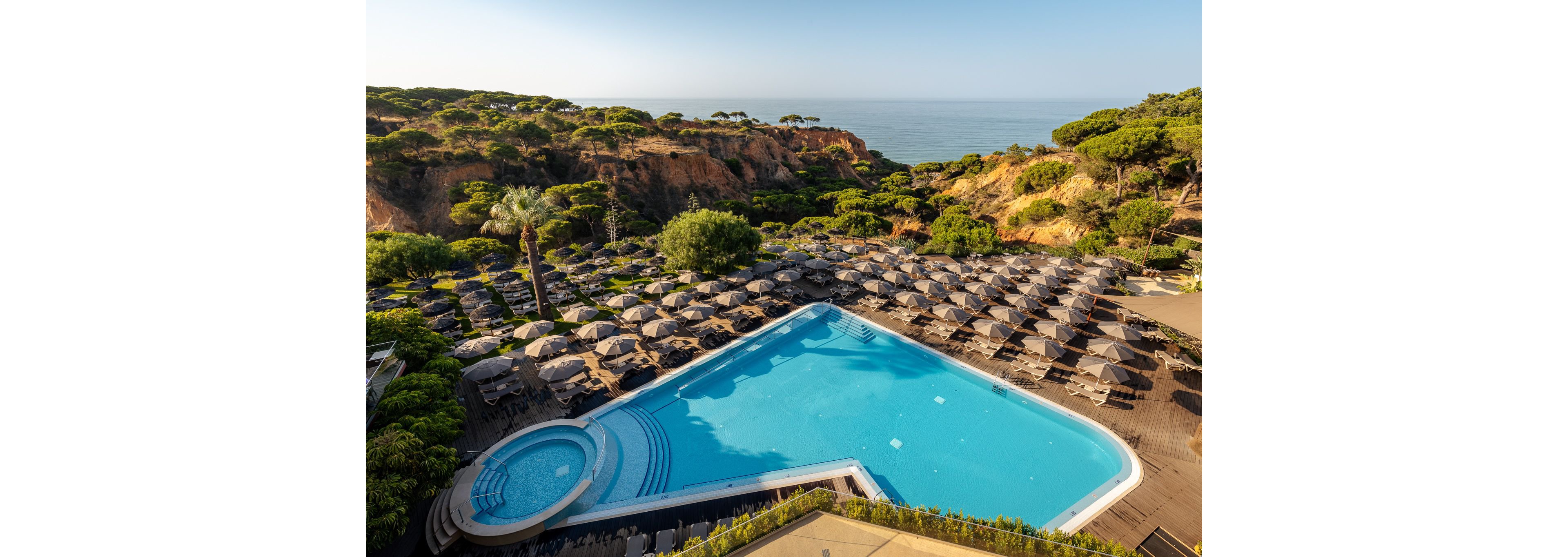 A large outdoor swimming pool sits on a clifftop resort surrounded by rows of sun loungers and umbrellas. Lush green trees and rocky cliffs frame the scene, leading the eye toward the calm ocean in the background. The pool area includes a smaller circular pool section and clean tiled deck, suggesting a relaxing holiday destination. Warm sunlight and clear skies create a serene, inviting atmosphere for vacationers.