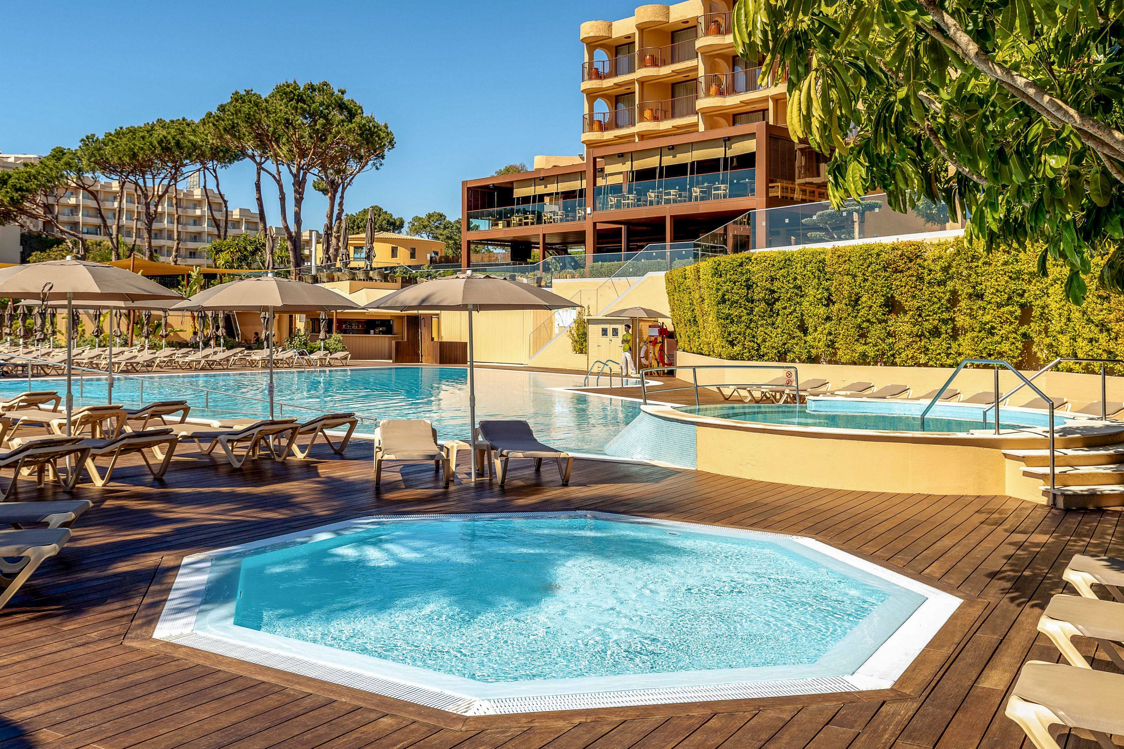 Outdoor resort pool complex featuring a small octagonal jacuzzi in the foreground. Sun loungers and beige umbrellas surround the pools on wooden decking, suggesting a relaxing vacation setting. Modern multi-story hotel buildings and tall trees rise in the background under a clear blue sky. The scene conveys a warm, sunny atmosphere ideal for leisure and summer holidays.