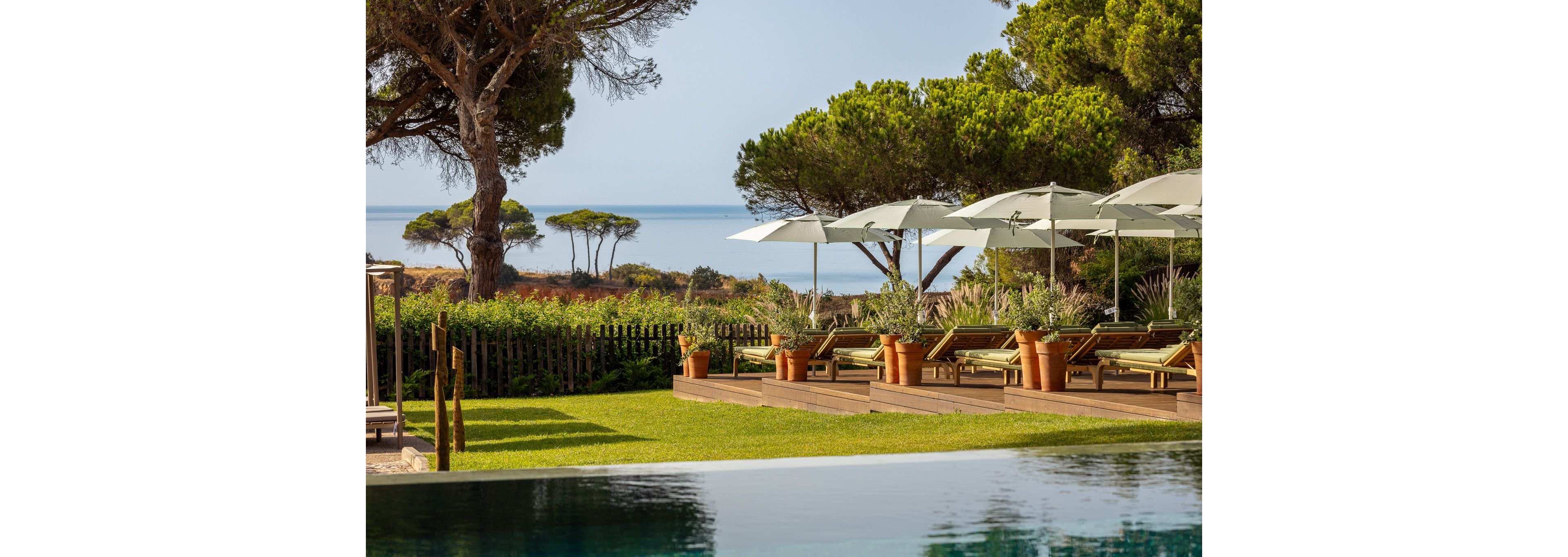 A serene outdoor pool terrace overlooks a calm sea framed by tall pine trees. Wooden sun loungers with white cushions are arranged under large white umbrellas on a raised deck. Lush green lawn and landscaped shrubs create a natural border between the pool area and the coastal view. The scene suggests a tranquil resort setting ideal for relaxation and sunbathing.