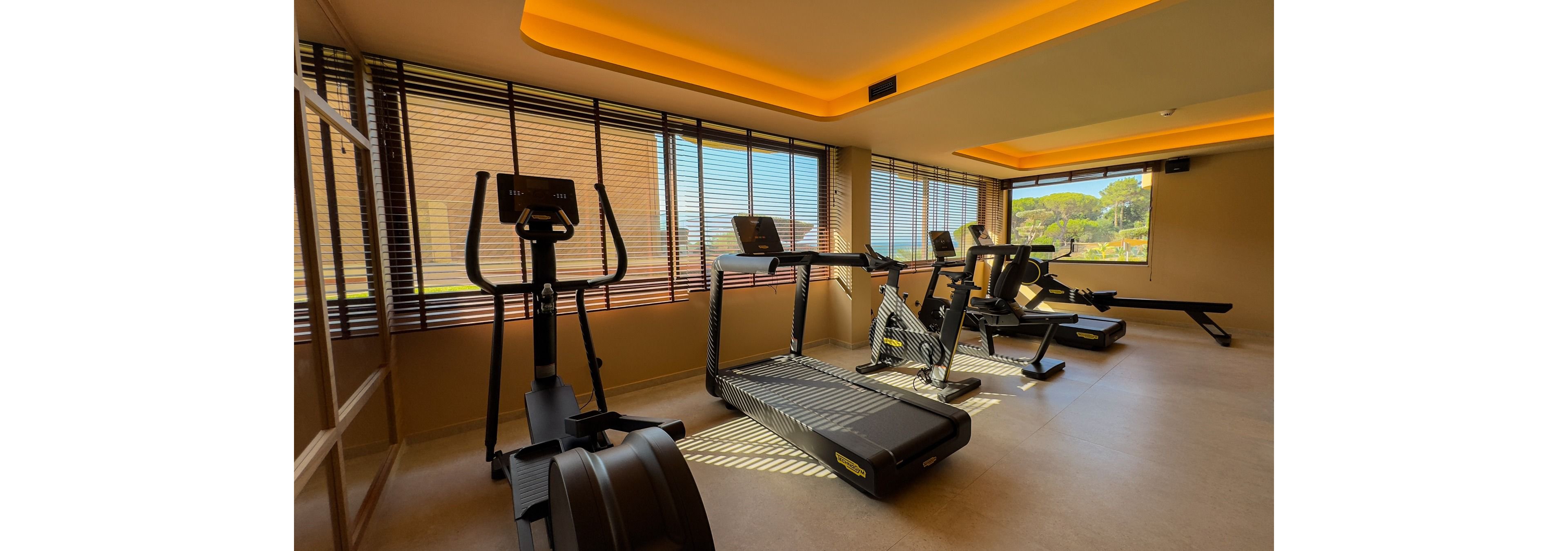A bright fitness room features several treadmills and an elliptical machine arranged in front of large windows. Warm ceiling lighting and beige walls create a comfortable, inviting workout atmosphere. Outside the windows, greenery and trees are visible, adding a natural view to the indoor gym setting. The space appears clean, modern, and ready for guests to exercise.
