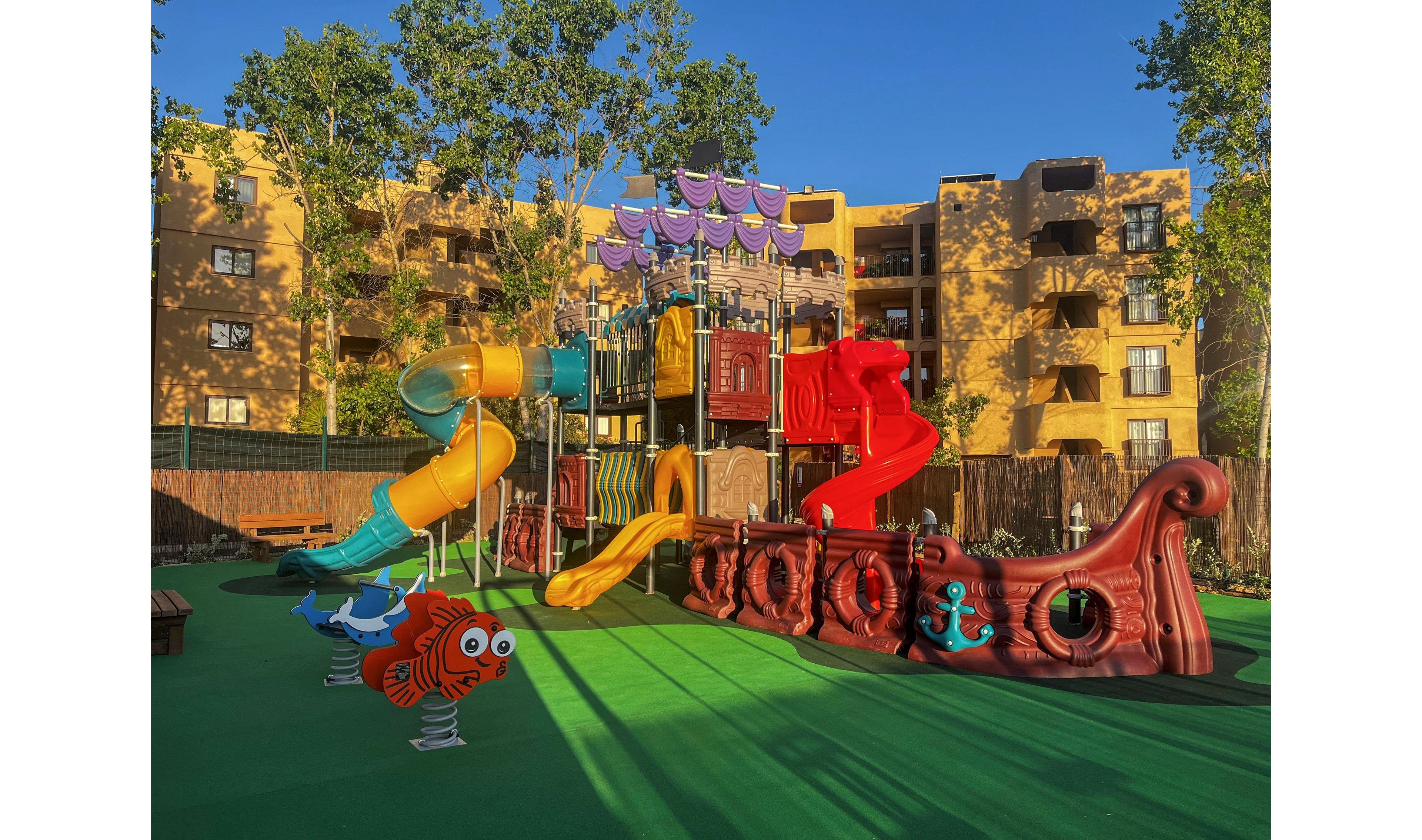 A vibrant pirate-themed playground structure stands in an outdoor courtyard under a clear blue sky. The play area features a large red ship with decorative anchors, multiple slides, and climbing elements in bright yellow, orange, and green. Spring-mounted fish characters and soft green safety flooring add to the playful atmosphere. Apartment-style buildings and tall trees surround the playground, creating an inviting family-friendly setting.