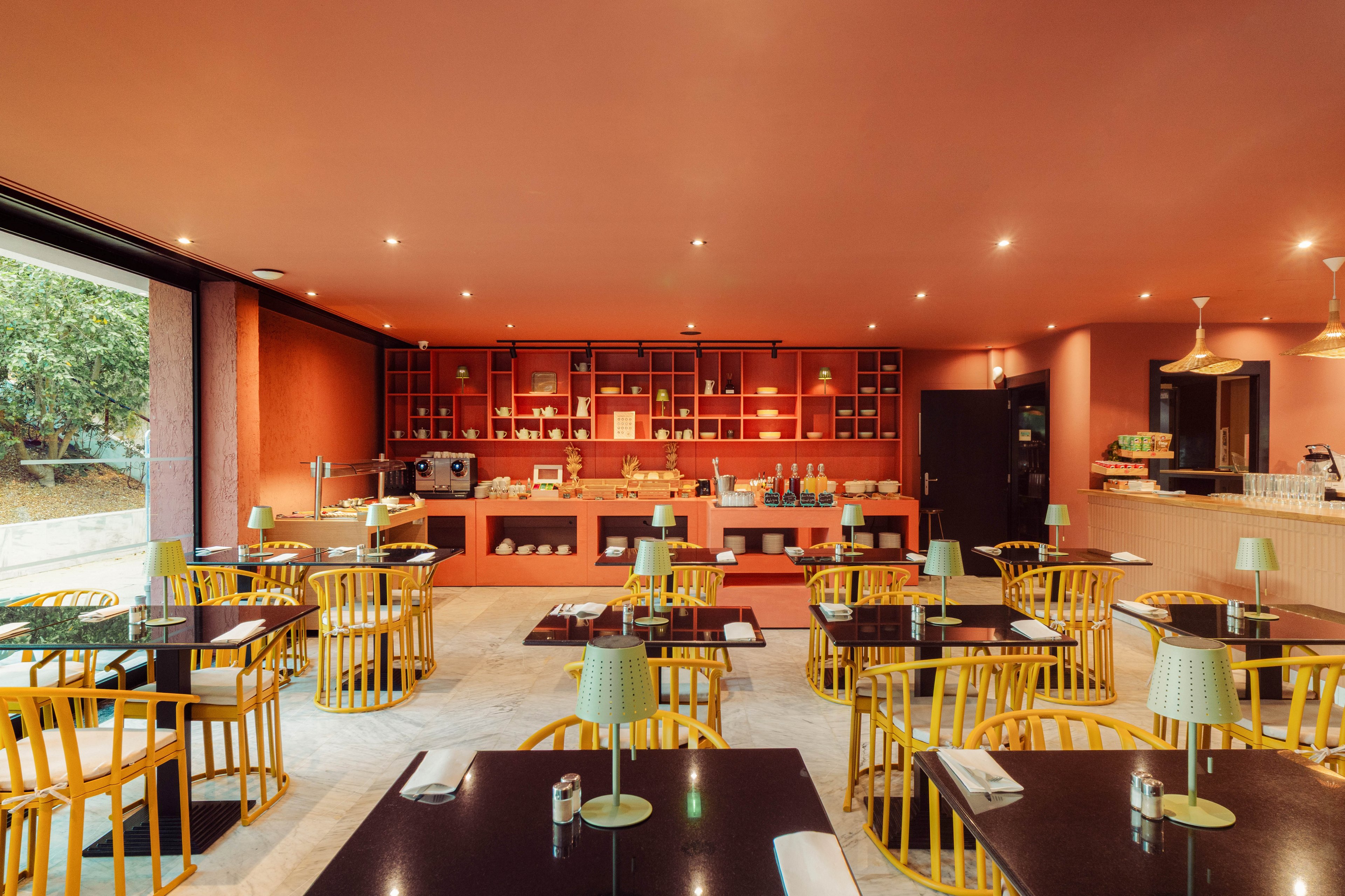 A contemporary cafe interior features neatly arranged tables and yellow chairs facing an open counter. The space has warm terracotta walls and ceiling, with recessed lighting creating a cozy atmosphere. Small table lamps with patterned shades sit on dark tabletops, adding a decorative touch. Shelving behind the counter displays various items, while large windows open to greenery outside.