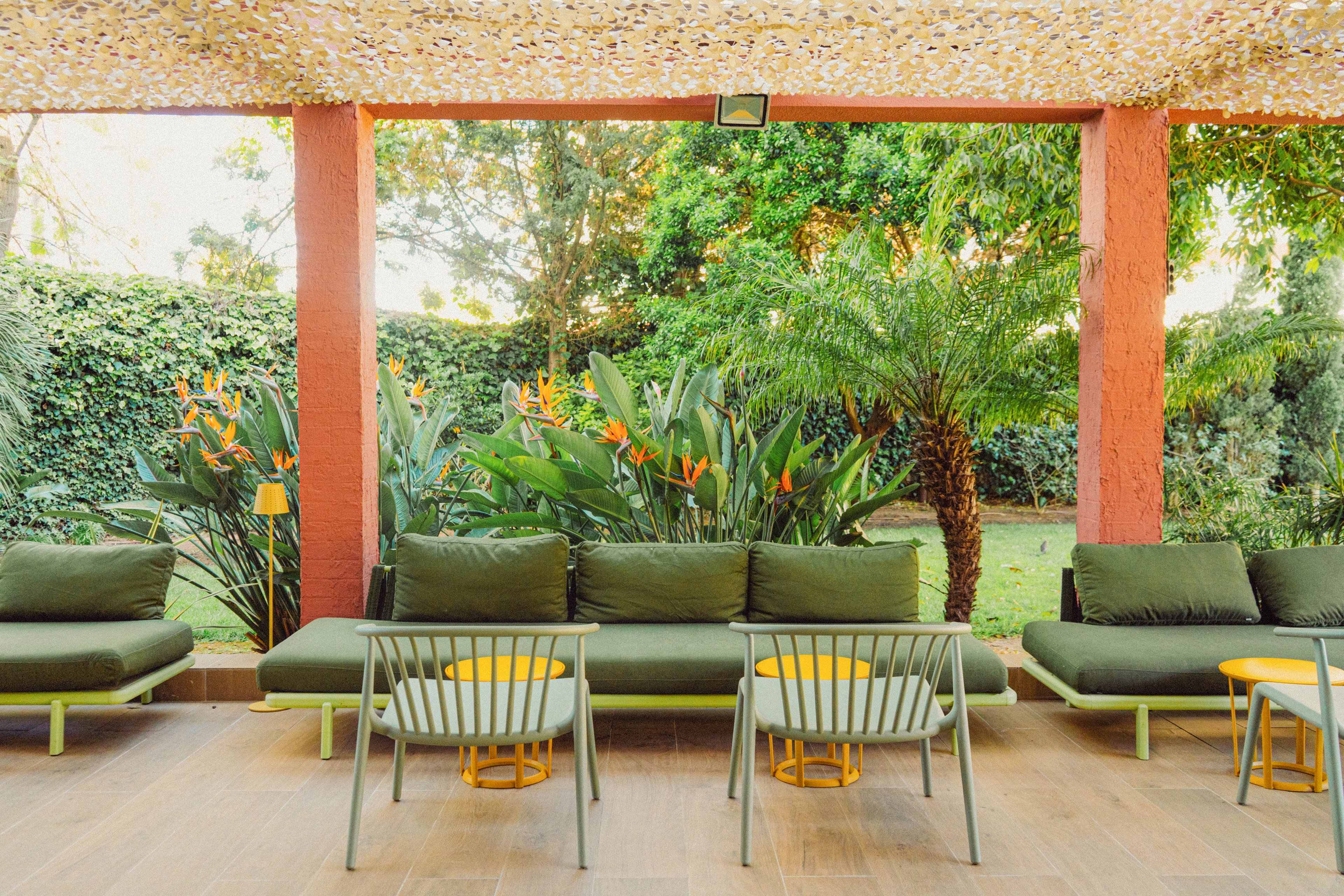 An inviting outdoor lounge area features long green cushioned sofas arranged under a shaded pergola. Simple metal chairs and small round tables with yellow tops create a relaxed seating arrangement. Lush tropical plants, including ferns and flowering shrubs, fill the background and frame the space. Warm wooden flooring and natural light contribute to a bright, tranquil garden atmosphere.