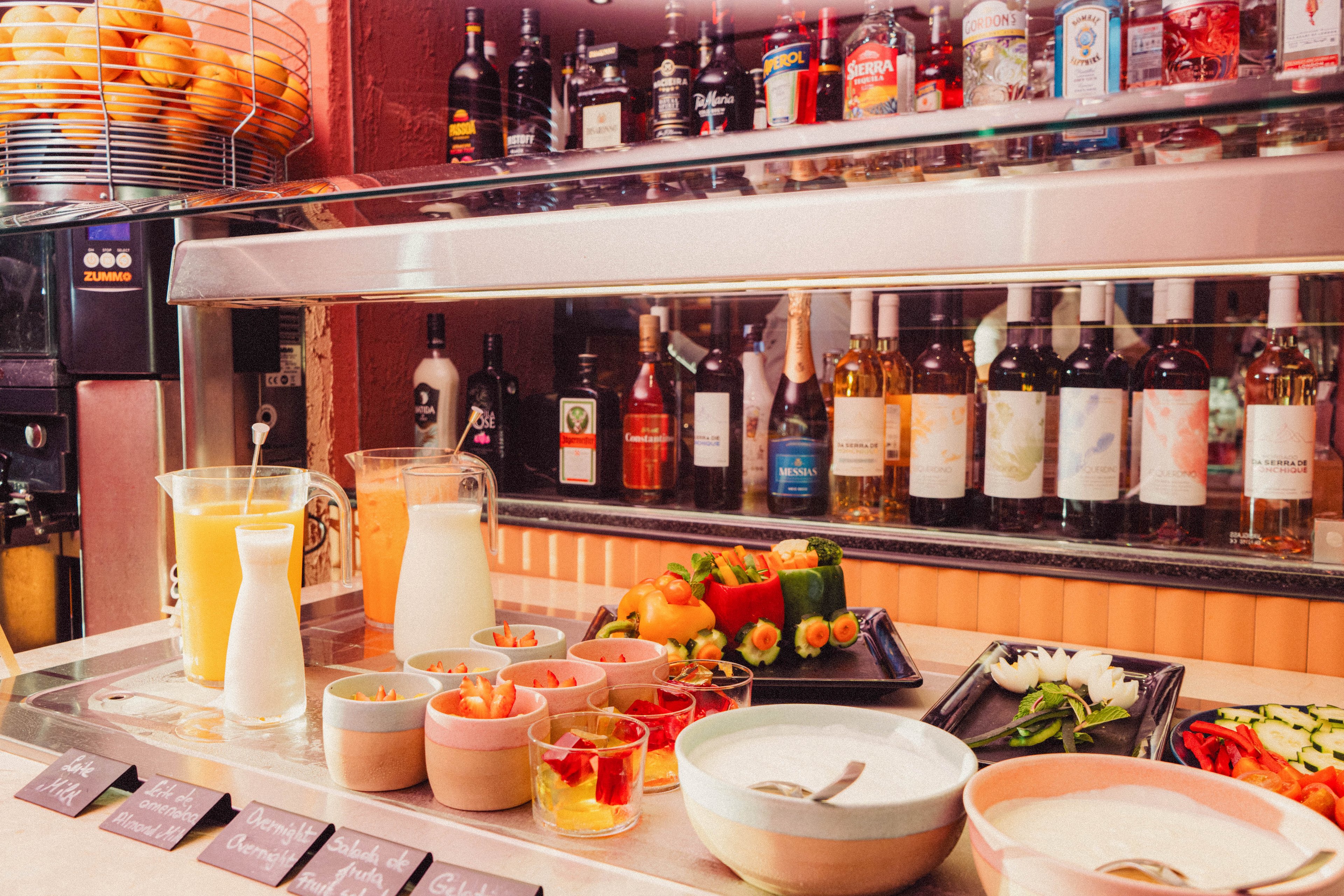 A breakfast buffet counter displays various bowls of yogurt, fruit, and small desserts in a brightly lit bar setting. Two large jugs of orange juice and milk stand beside pastel-colored bowls and glasses. Behind the counter, shelves are lined with assorted wine and liquor bottles, adding depth and color to the scene. Fresh vegetables and garnishes are arranged in trays, contributing to a vibrant and inviting atmosphere.