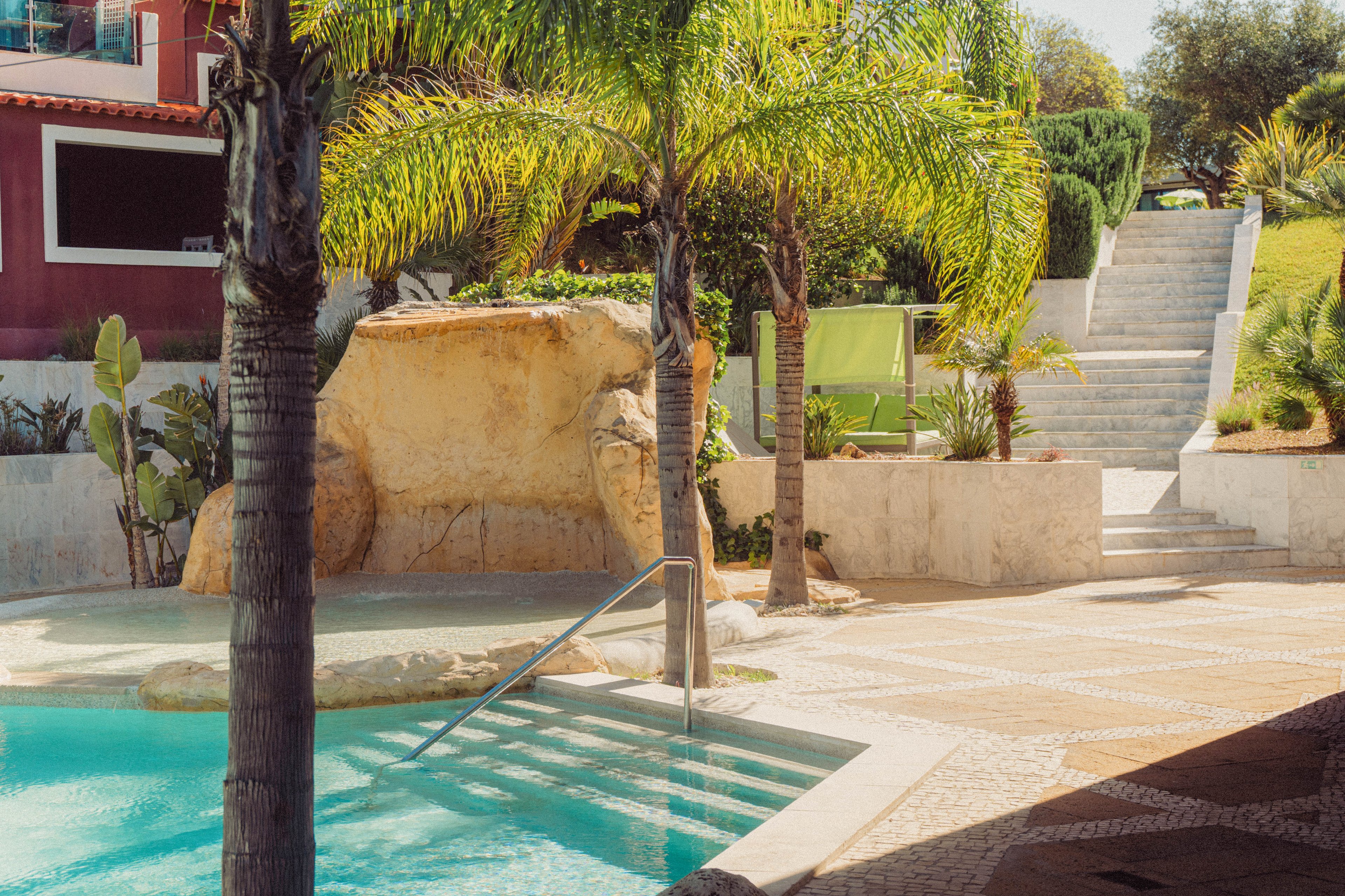 A bright outdoor swimming pool is surrounded by palm trees and stone features in a villa setting. A rock formation and tiled pool deck create a resort-like atmosphere. In the background, a staircase leads up through a landscaped garden with trimmed shrubs and grass. Warm sunlight enhances the tropical feel of the scene, highlighting the turquoise water and light stone surfaces.