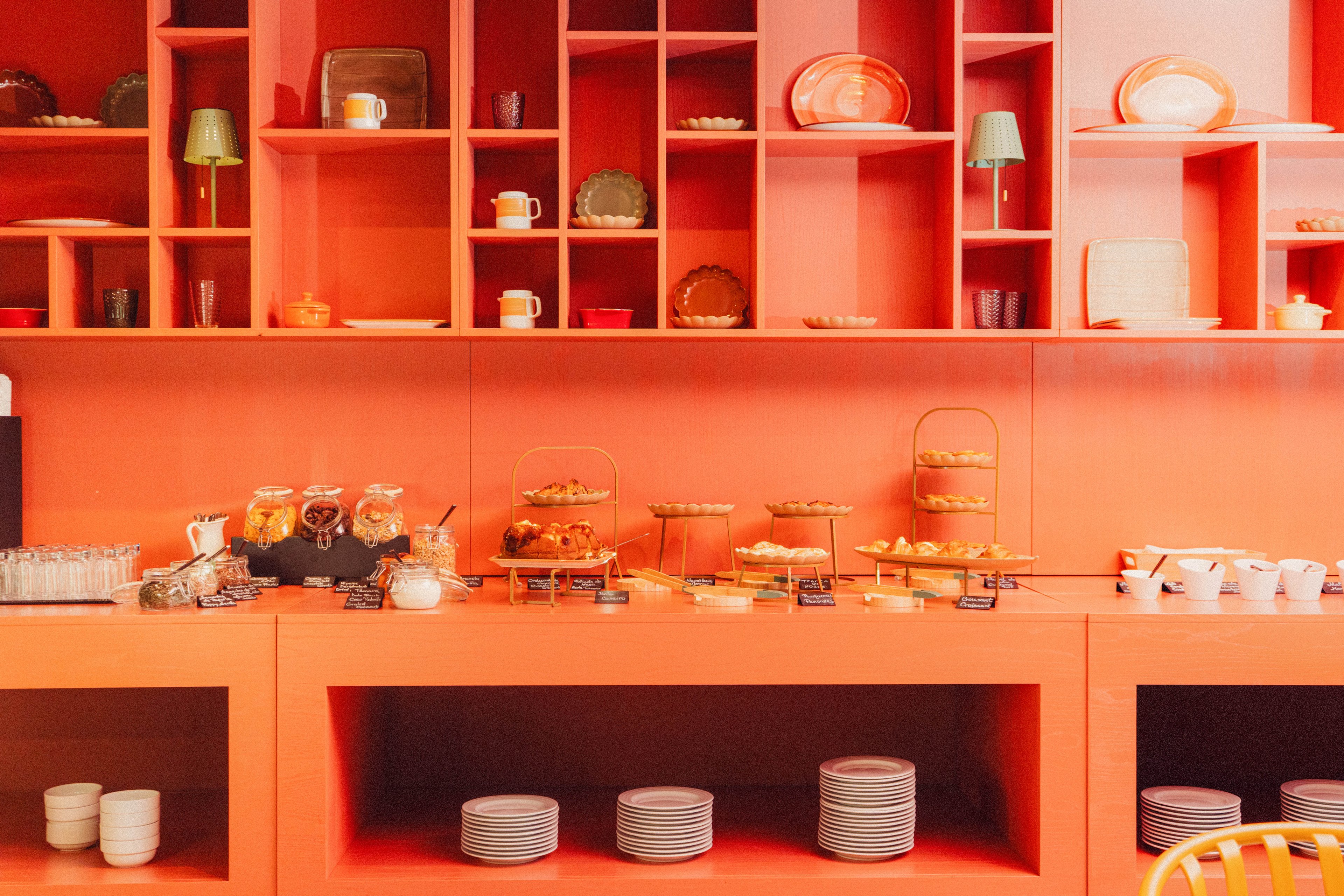 A contemporary buffet counter displays an assortment of pastries and baked goods arranged on tiered stands. The setting features a bold orange wall with open shelving filled with plates, cups, glasses, and decorative objects. White dishes and glassware are neatly stacked in cubbies below the counter, emphasizing order and symmetry. Warm lighting and the monochromatic orange palette create a cozy yet stylish café or hotel breakfast atmosphere.