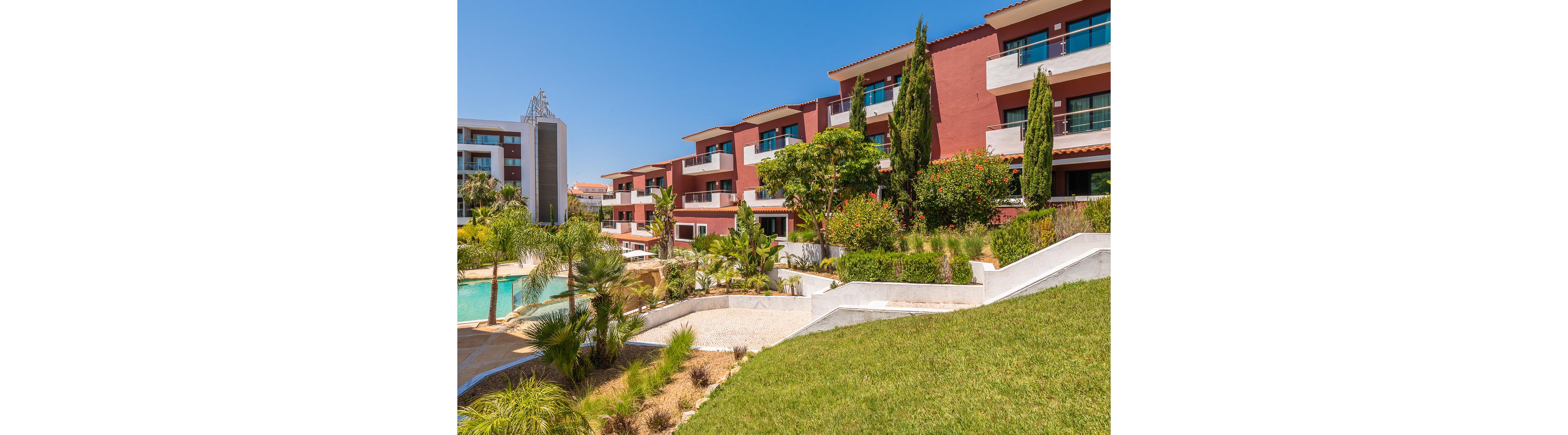 A contemporary apartment complex features red and white facades overlooking a lush landscaped courtyard. The setting includes a winding swimming pool bordered by palm trees, shrubs, and manicured lawns. Bright sunlight and a clear blue sky create a warm, inviting atmosphere. Multiple balconies and terraces suggest residential or vacation accommodations in a resort-like environment.