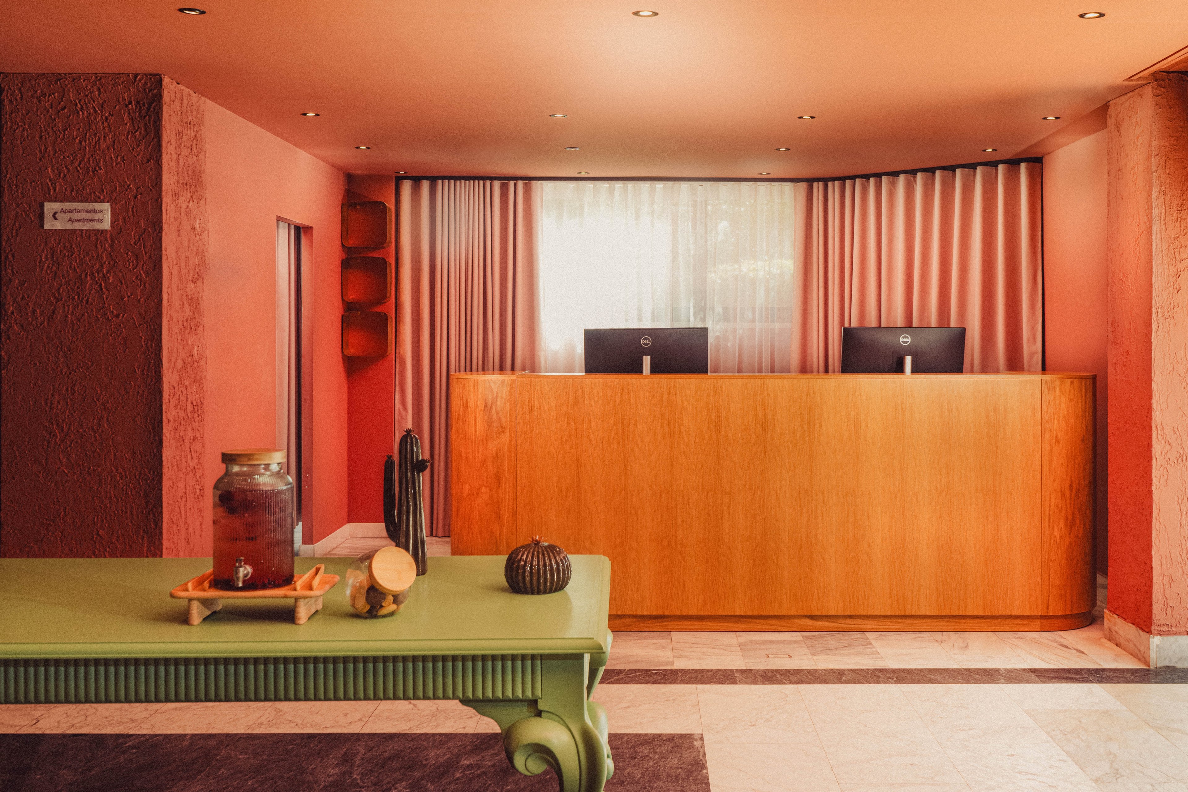 A modern hotel reception desk stands against a warm pink wall with soft curtains and two computer monitors. In the foreground, a green table holds a glass beverage dispenser, a small cactus, and decorative objects. Recessed ceiling lights illuminate the minimalist lobby, highlighting the contrast between the wooden counter and colorful walls. The overall scene feels welcoming and contemporary with a retro-inspired palette.