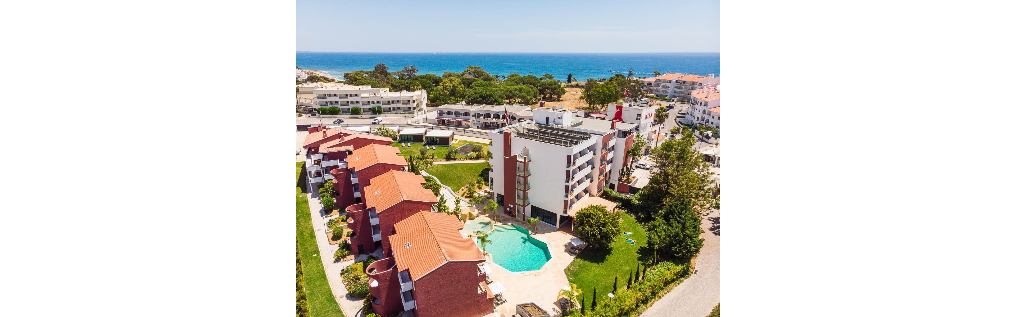 An aerial view shows a modern coastal resort complex with multiple red-roofed buildings and a central swimming pool. The property is surrounded by green lawns, trees, and landscaped gardens, creating a relaxing vacation atmosphere. In the background, the blue ocean and sandy shoreline stretch along the horizon under a clear sky. Nearby residential buildings and roads indicate the resort is located in a developed seaside town.