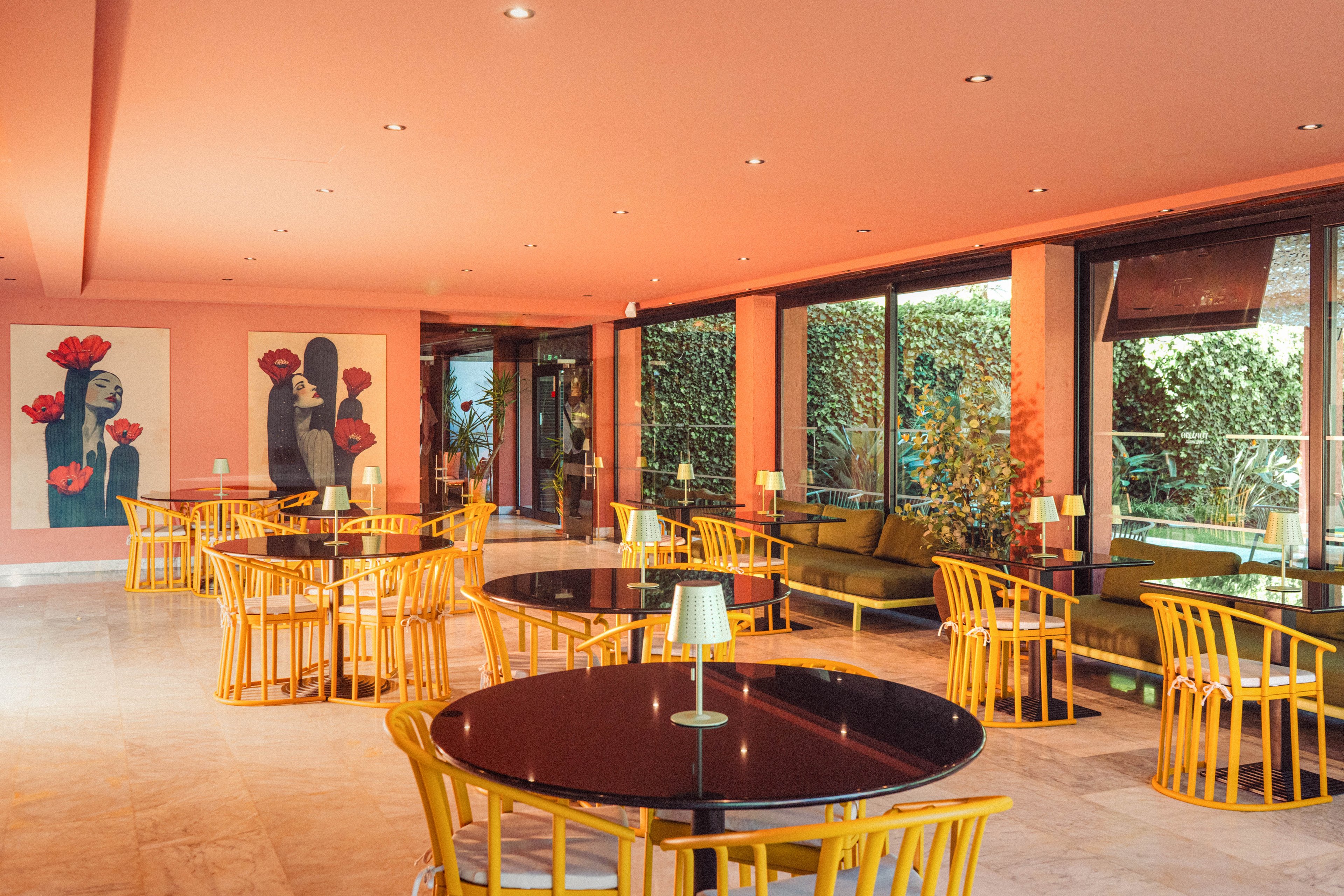 A contemporary lounge area features round black tables paired with bright yellow chairs. The room has pink walls and ceiling, large windows, and glass doors opening to a green outdoor hedge. Wall art with stylized portraits and red flowers adds a decorative focal point. Soft lighting from ceiling spots and small table lamps creates a warm, inviting atmosphere. The space appears calm and ready for guests in a hotel, café, or restaurant setting.