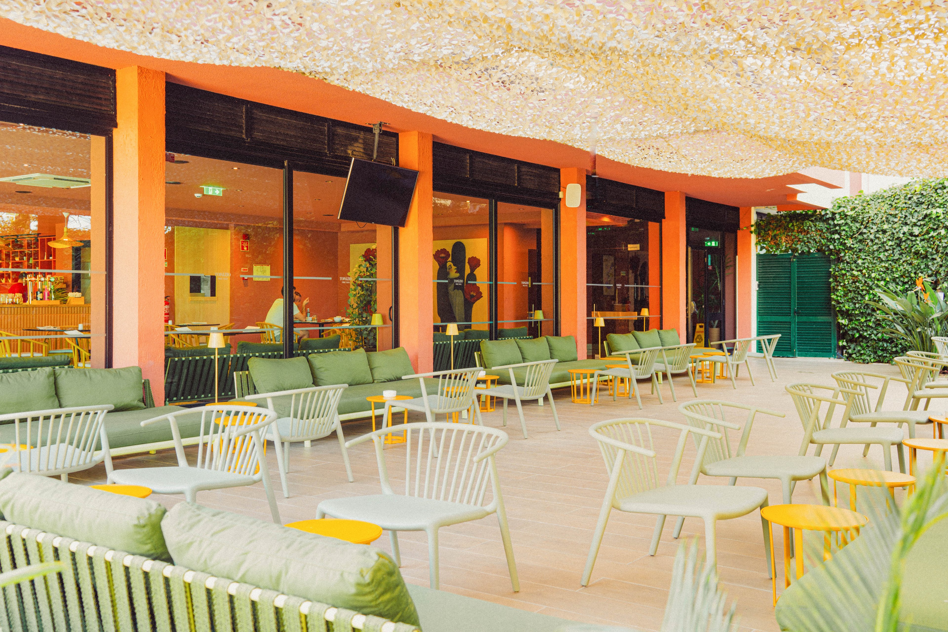 An outdoor lounge area features rows of modern metal chairs and cushioned sofas arranged around small round tables. The space is shaded by a light, textured canopy and bordered by a hedge and colorful building facade. Large windows reveal an indoor bar area with warm lighting and bottles on display. The overall scene feels relaxed and inviting, suitable for socializing or casual dining.