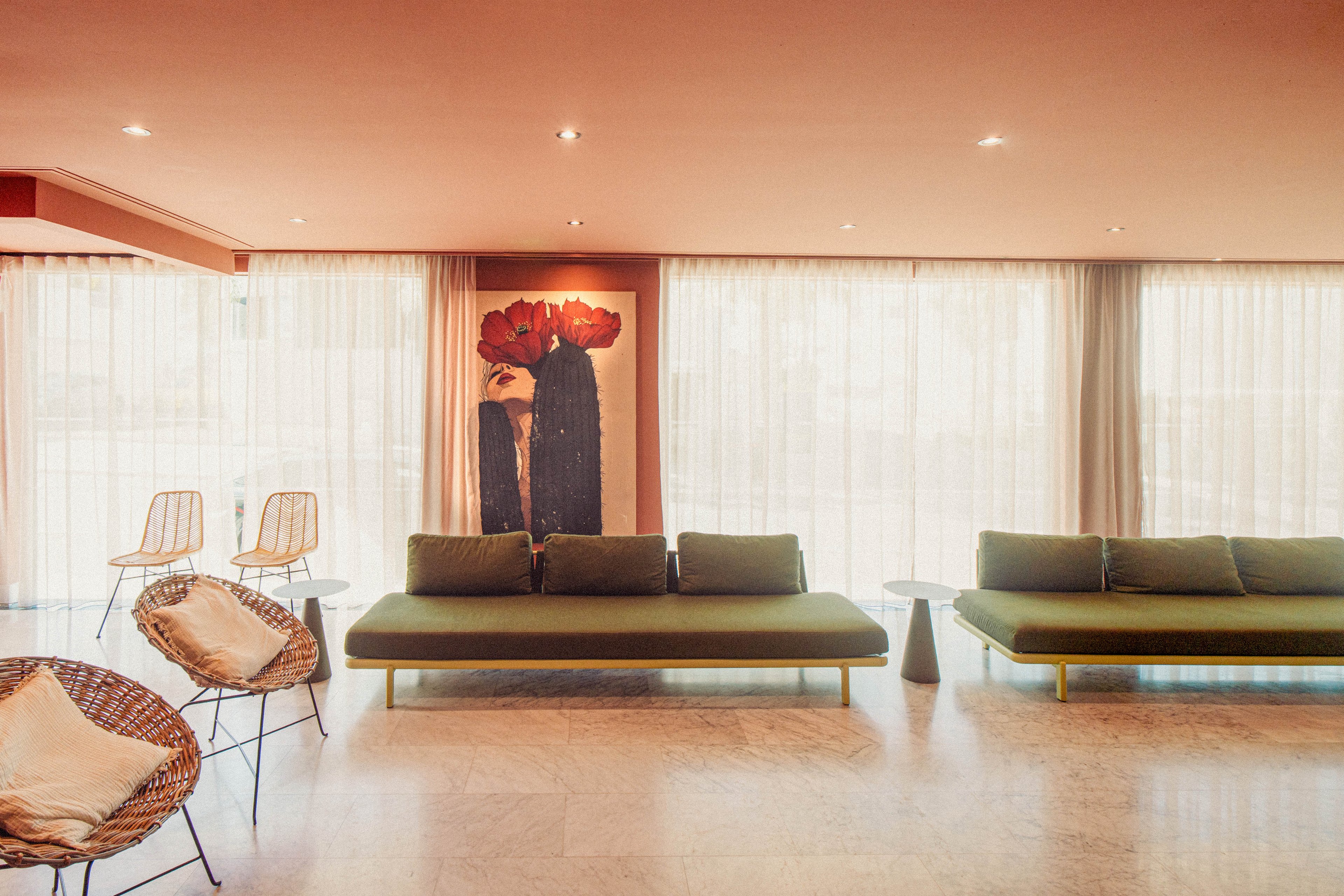 A contemporary lounge features long green sofas arranged against a wall of sheer floor-to-ceiling curtains. Warm lighting from recessed ceiling fixtures creates a cozy, inviting atmosphere. Wicker chairs with cushions and slim metal barstools add texture and variety to the seating options. A large artwork with bold red flowers provides a striking focal point in the otherwise minimalist space.
