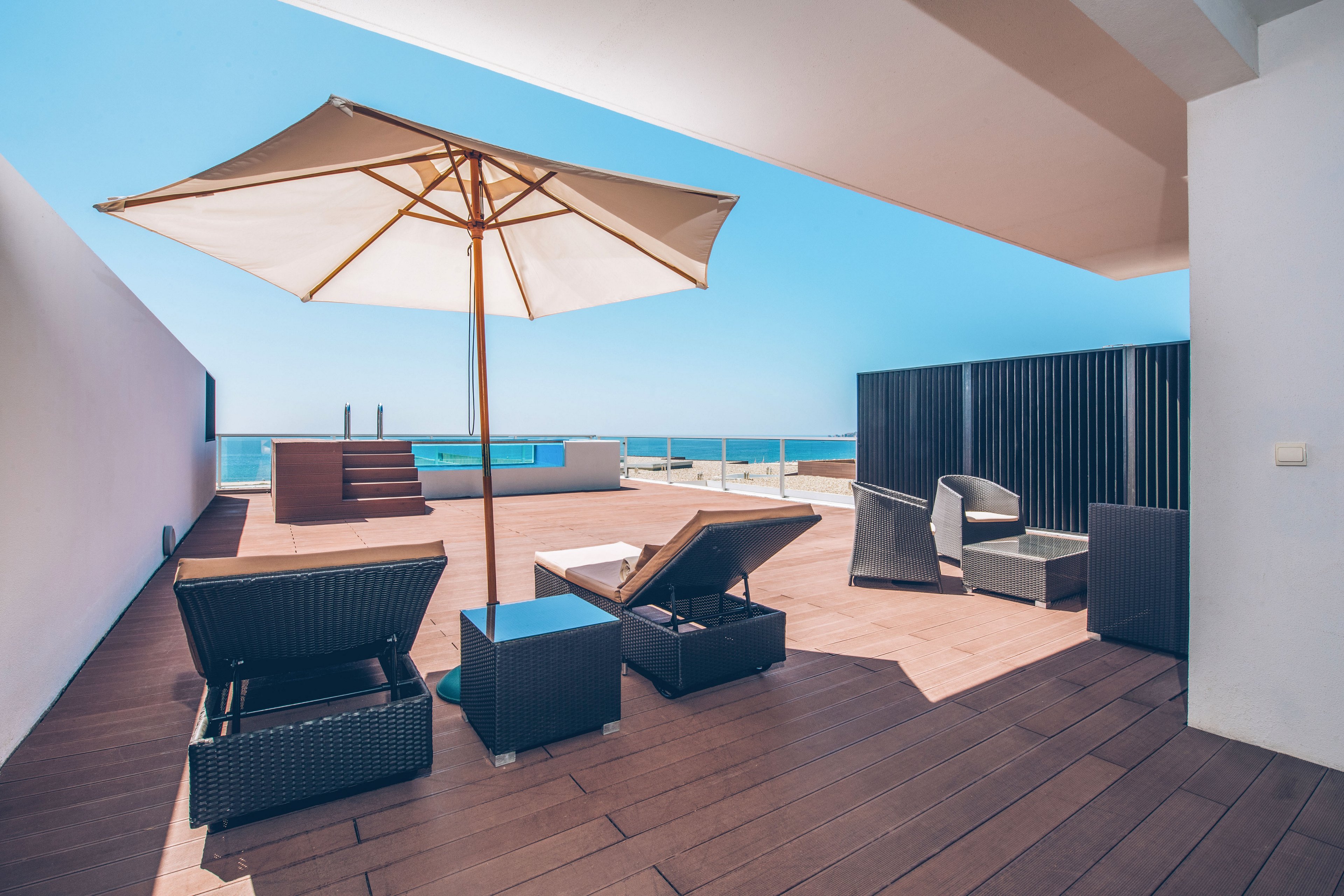A contemporary rooftop terrace features sun loungers and wicker chairs arranged on a wooden deck. A large beige umbrella provides shade in the center of the outdoor space. Glass railings frame a clear view of the blue ocean and sky in the background. Clean architectural lines and minimal decor create a relaxed, luxurious coastal atmosphere.