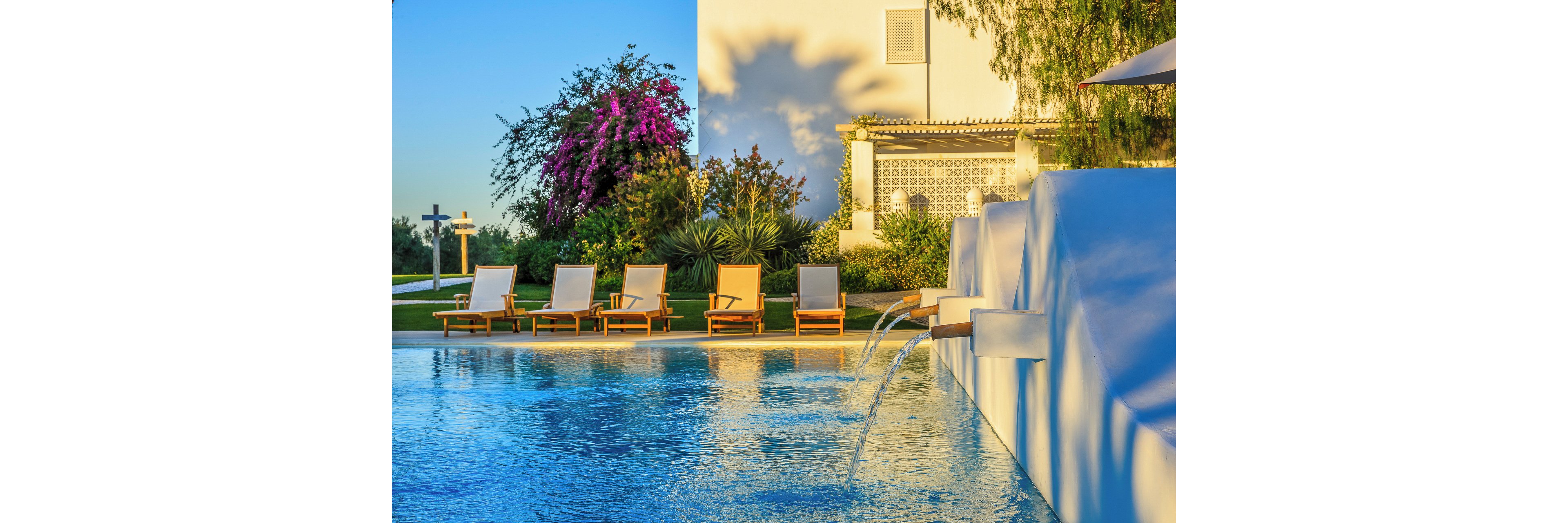 A tranquil outdoor swimming pool glows under warm sunlight beside a white building. Wooden lounge chairs with cushions line the poolside, facing the clear blue water. Small fountains cascade into the pool, creating gentle ripples. Lush greenery and vibrant flowering plants add color and a relaxed vacation atmosphere to the scene.