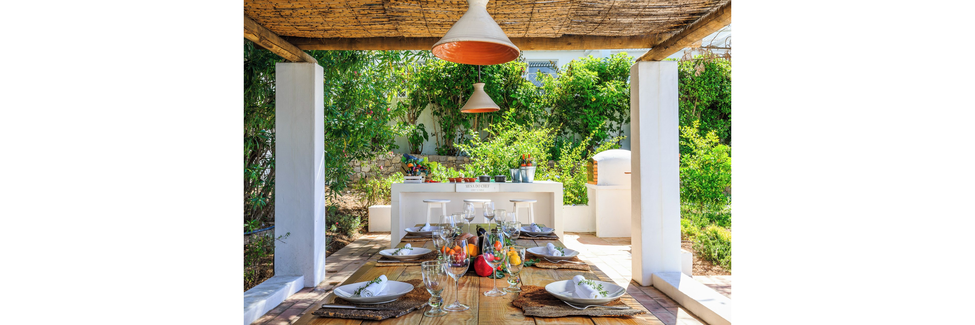 A rustic wooden table is elegantly set for a meal under a shaded pergola in a lush garden. Glassware, white plates, and folded napkins are arranged neatly alongside fresh fruit and vegetables as a centerpiece. In the background, a white outdoor counter with stools and hanging pendant lights creates an inviting cooking and serving area. Green foliage and natural materials give the scene a relaxed Mediterranean atmosphere.